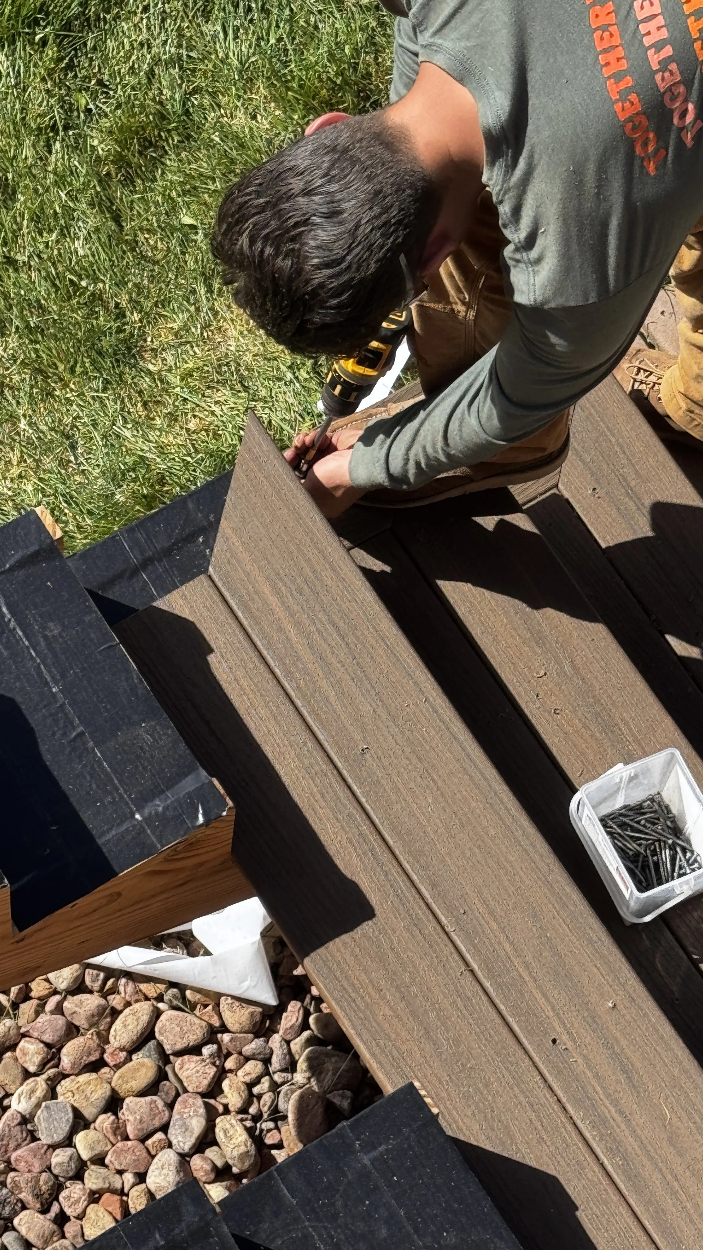 A person wearing a gray shirt and work pants using a power drill to secure a wooden plank to a structure outdoors. There are rocks on the ground and a small plastic container filled with screws on the wooden surface.