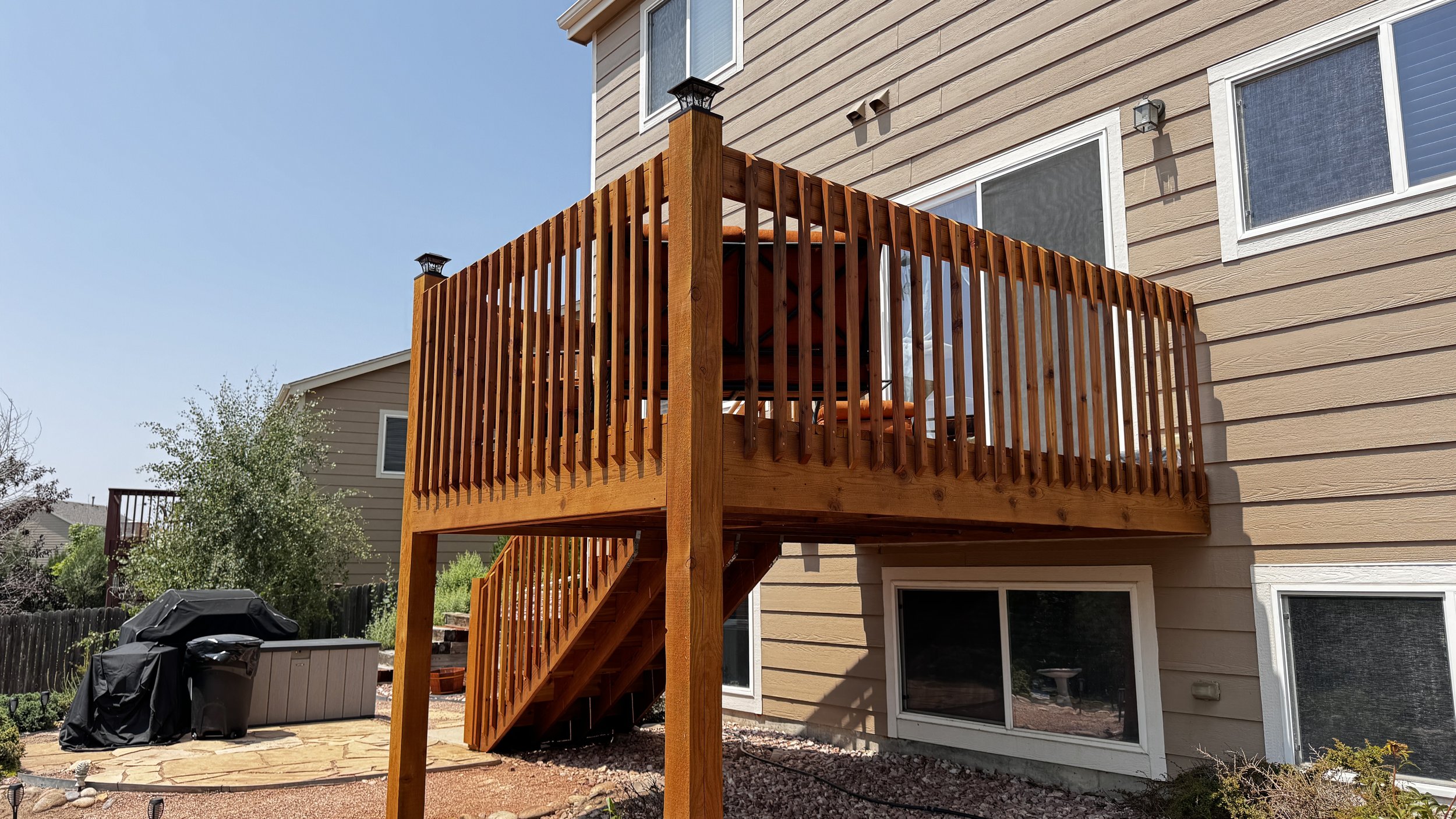 Backyard view of a two-story house with beige siding, showing a wooden deck with stairs and a railing, some outdoor furniture, a covered grill, and a garden area with trees and bushes under a clear blue sky.