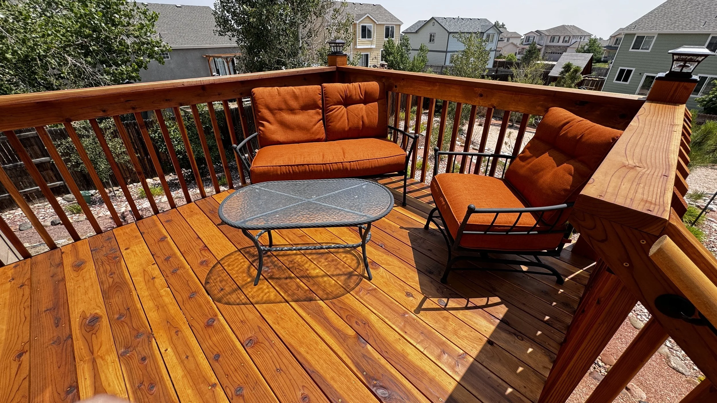 A wooden balcony with two orange cushioned chairs, a small glass table, and surrounding houses in a suburban neighborhood.