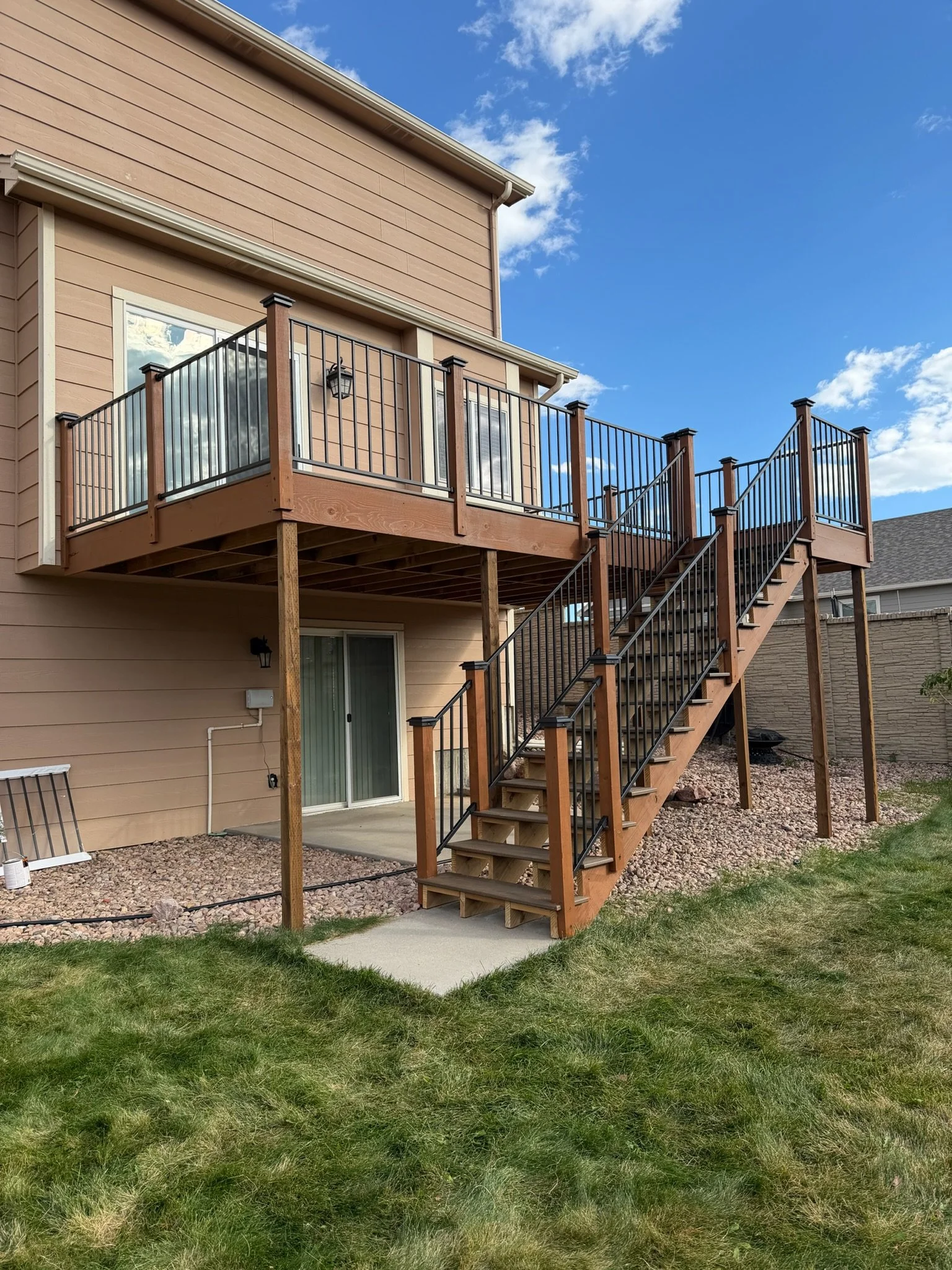 A two-story house with a wooden deck and black metal railing outside, accessible by stairs, in a suburban backyard with a green lawn and a gravel area.