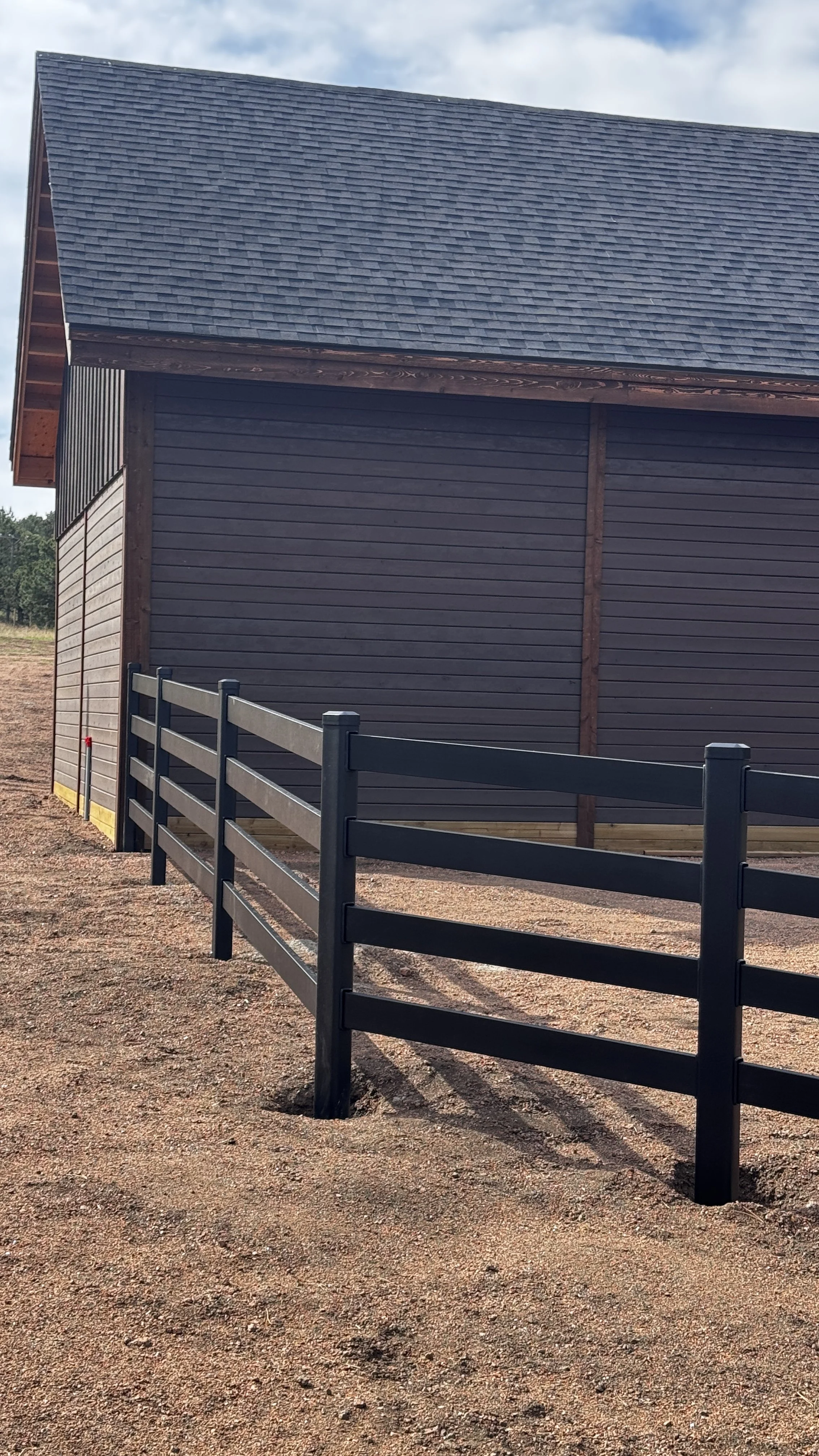 A wooden barn with dark horizontal siding, a metal roof, and a black fence in front, on a dirt ground with a cloudy sky above.