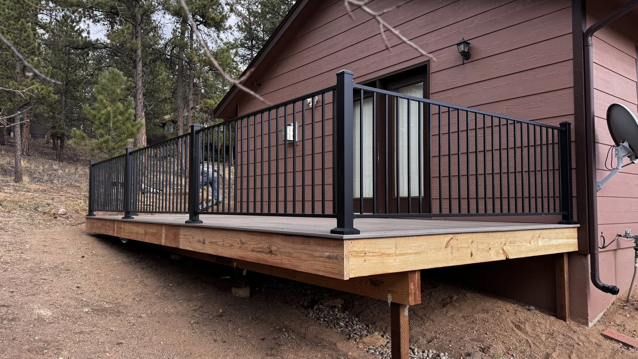 A small wooden deck with black metal railing attached to a reddish-brown house, surrounded by trees. There's a satellite dish attached to the house and some outdoor equipment visible.