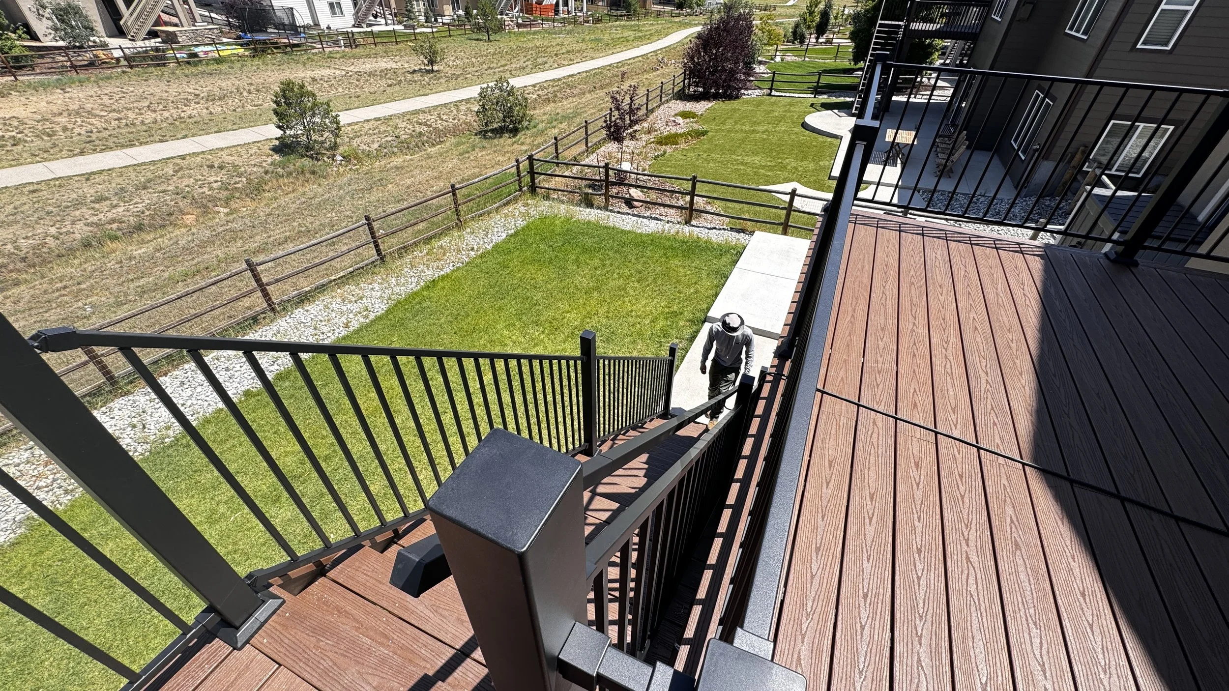 A view from an apartment balcony overlooking a small, fenced grassy yard with a person walking down the stairs. The yard has some trees and a sidewalk running through a nearby open area.
