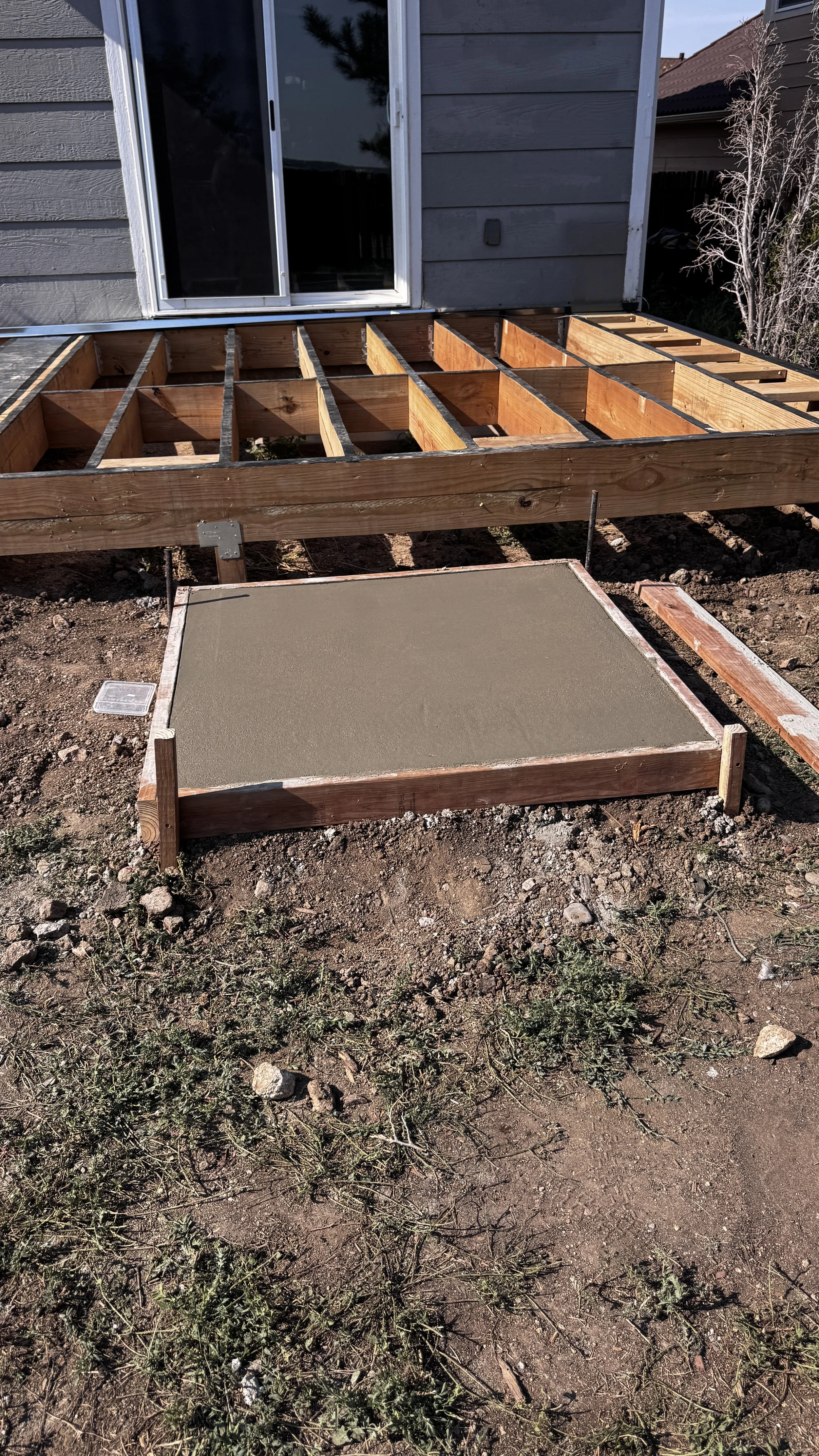 Wooden frame for a deck or patio under construction, with a gray concrete slab on the ground, in front of a house with gray siding and a sliding glass door.