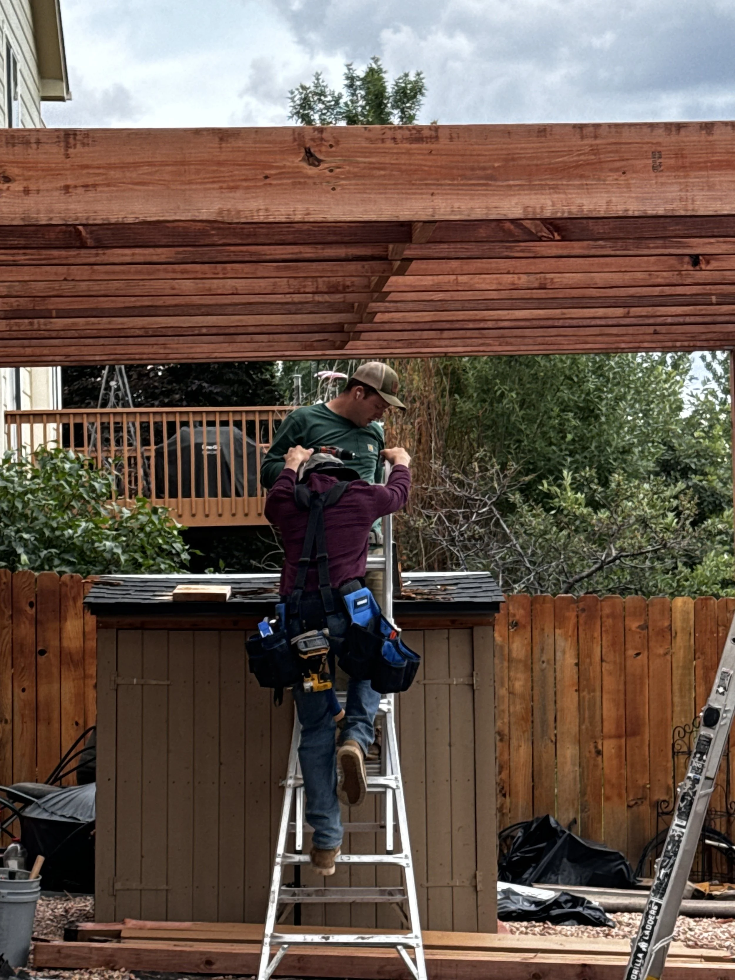 A man on a ladder installing or repairing a wooden roof over a backyard with a wooden fence and trees, with tools and equipment nearby.