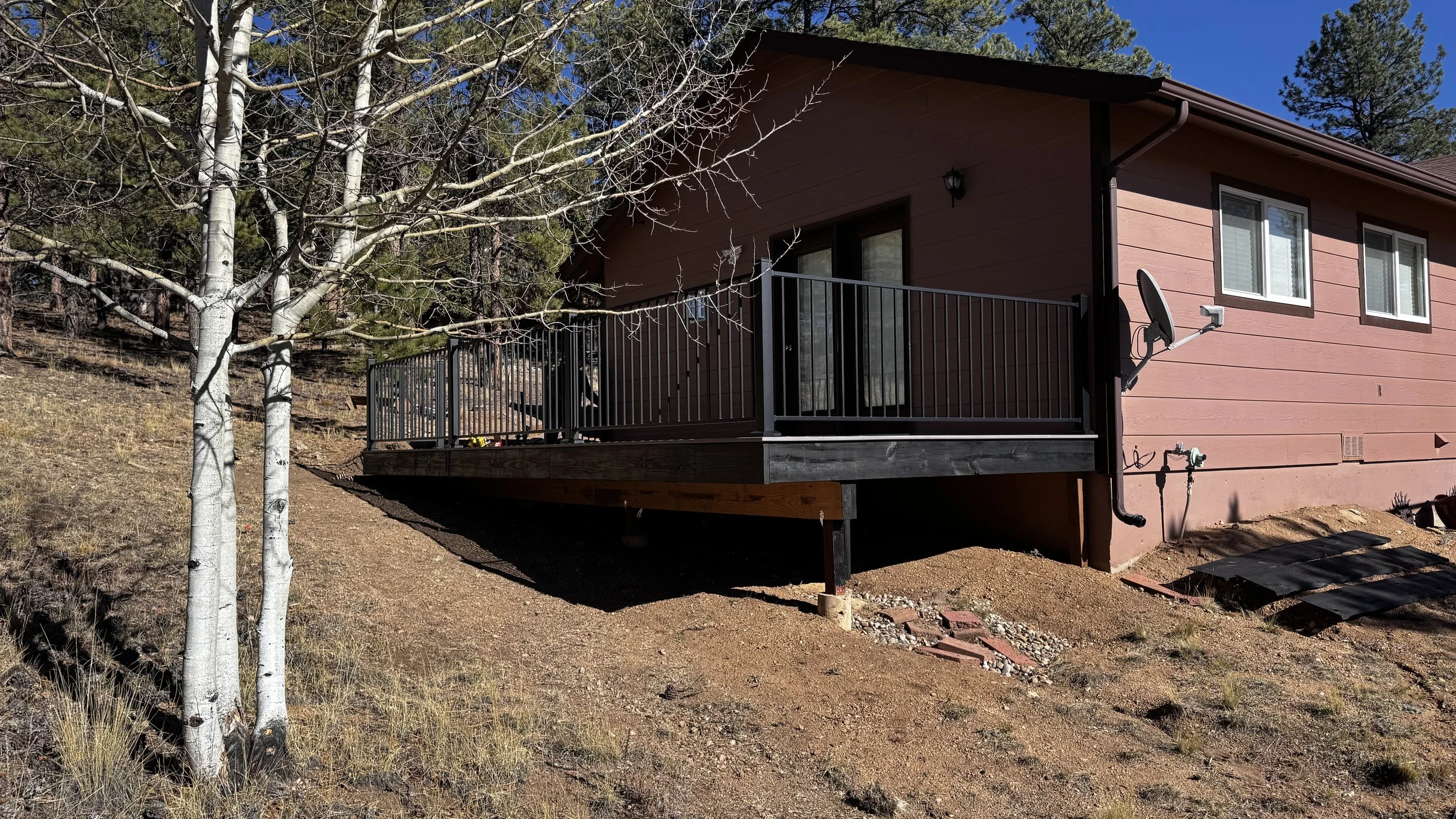 The side of a pink house with a small balcony and three windows, surrounded by tall trees with bare branches, on a dirt and gravel yard under a clear blue sky.