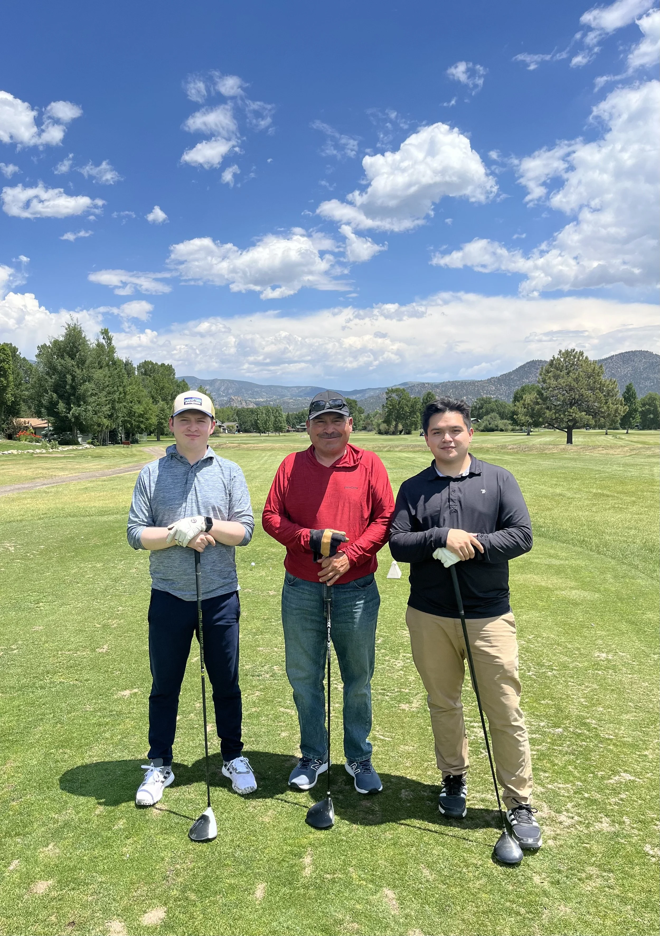 Three men standing on a golf course holding golf clubs, with trees, mountains, and a blue sky with white clouds in the background.