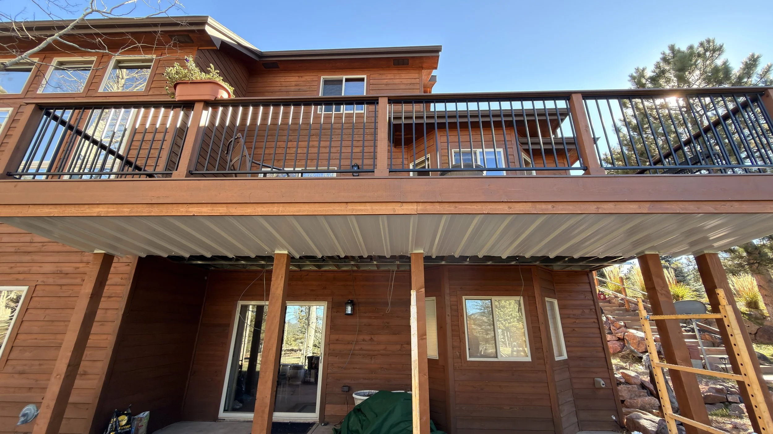 A two-story wooden house with a deck on the upper level, black metal railing, and large windows. The lower level is under construction with wooden support beams and an open area.