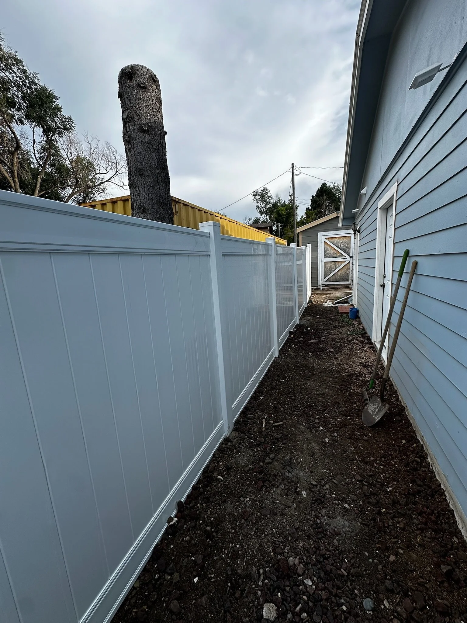 Backyard with white vinyl fence, blue house, dirt ground, garden tools leaning against the house, large tree trunk, cloudy sky.