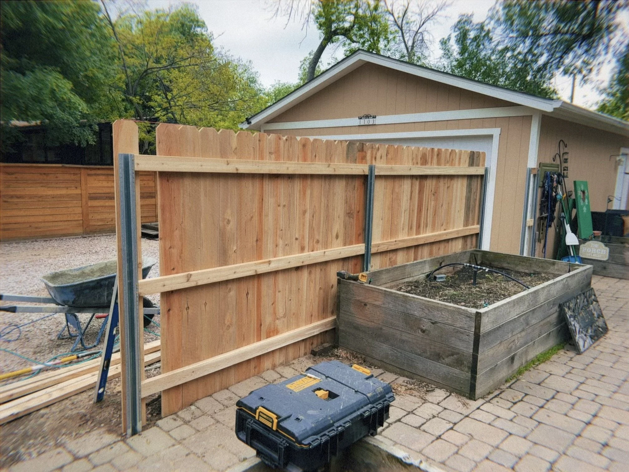 A backyard is under construction with a new wooden fence being built. Construction tools and a toolbox are on the ground. There is a wheelbarrow, and a raised garden bed with soil. The house is in the background with trees overhead.