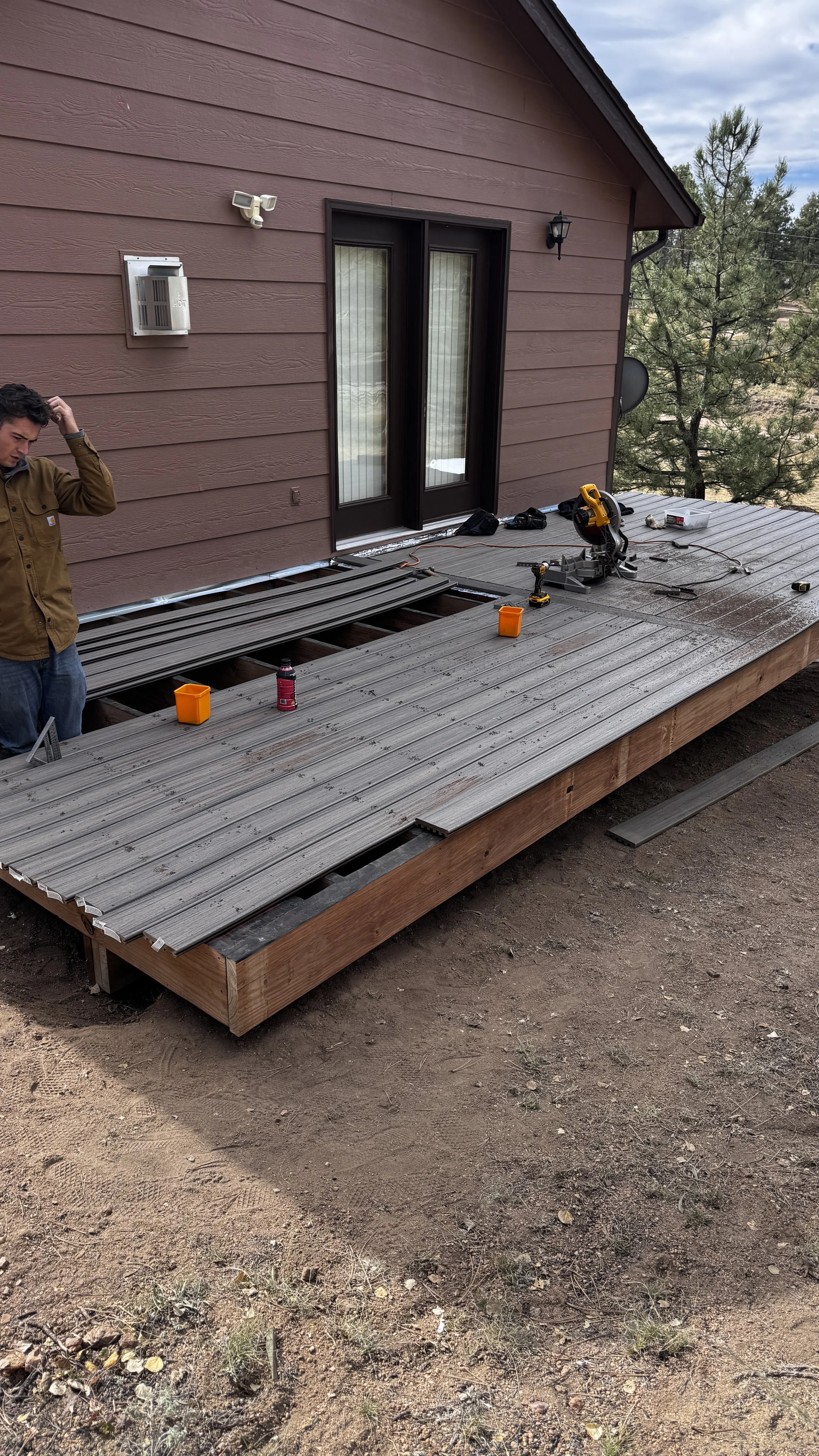 A person standing on a wooden deck under construction outside a house, with tools and materials like a drill, saw, and screws laid out on the deck. The house has red siding and has a sliding glass door, outdoor light fixtures, and a sky with some clo