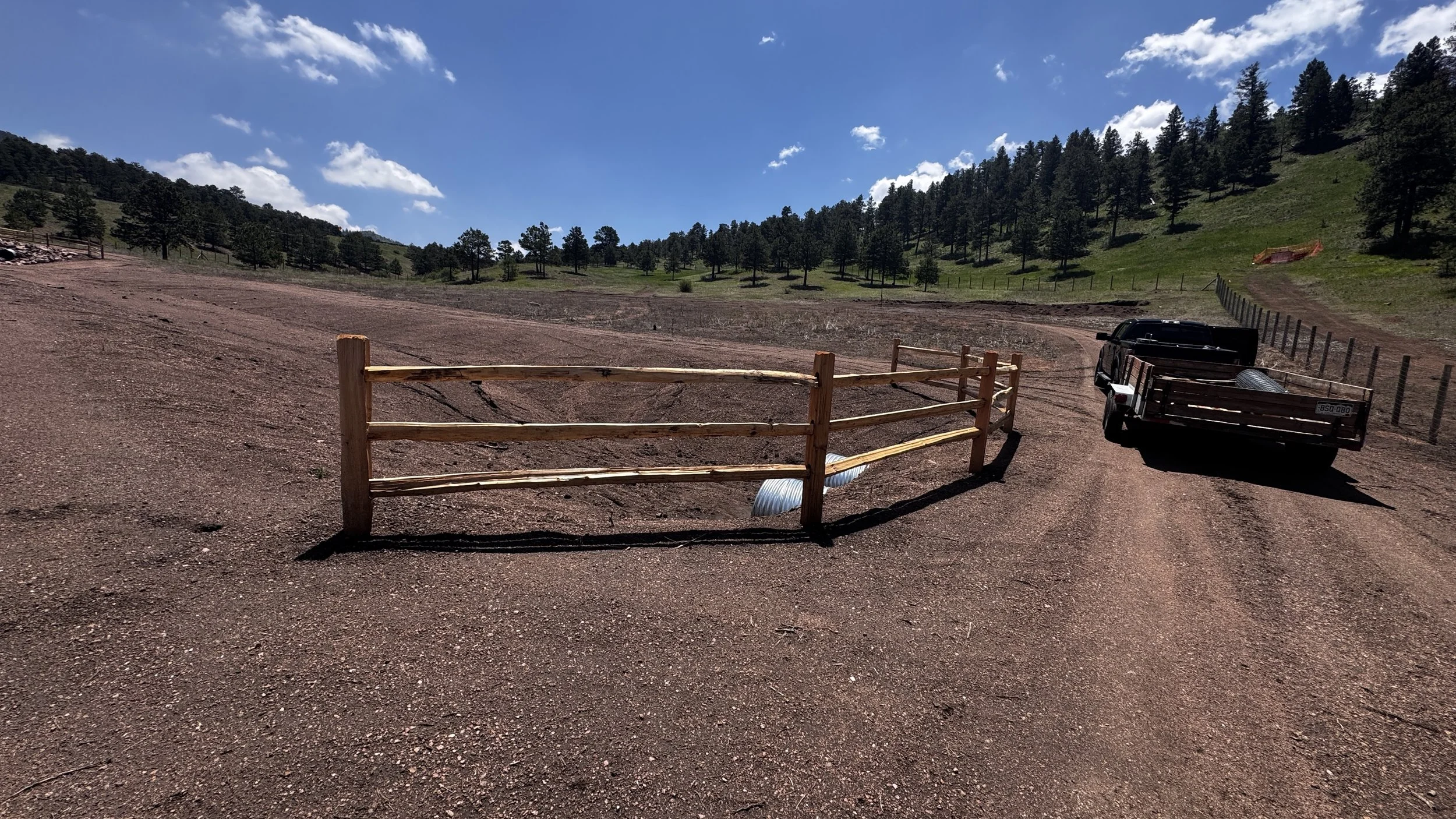 A dirt road with a wooden fence on the left and a trailer on the right side. The area is surrounded by mountains and trees with a blue sky and scattered white clouds.