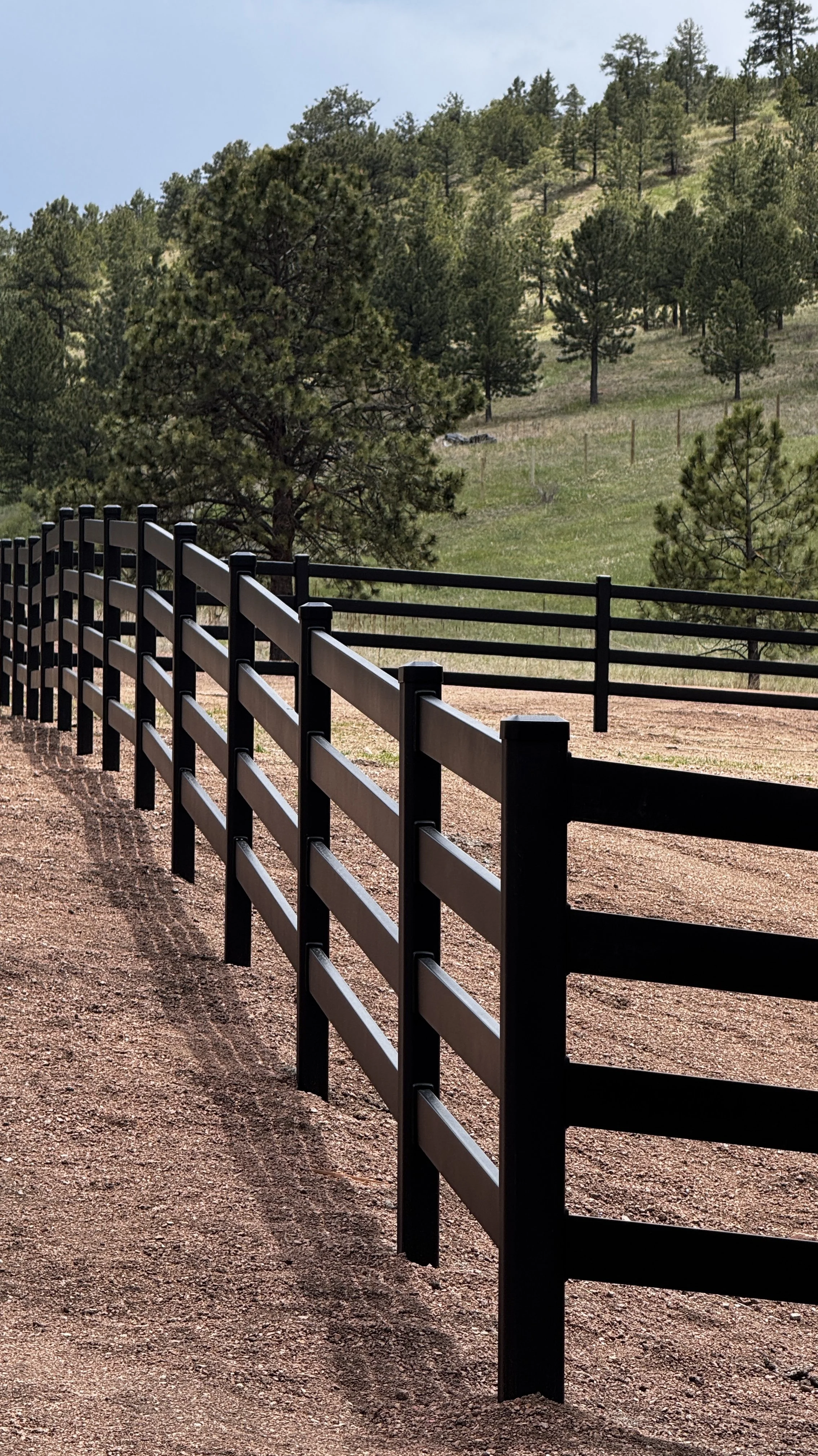 A black buckly steel horse fence running along a dirt road with a background of green trees and a hillside.