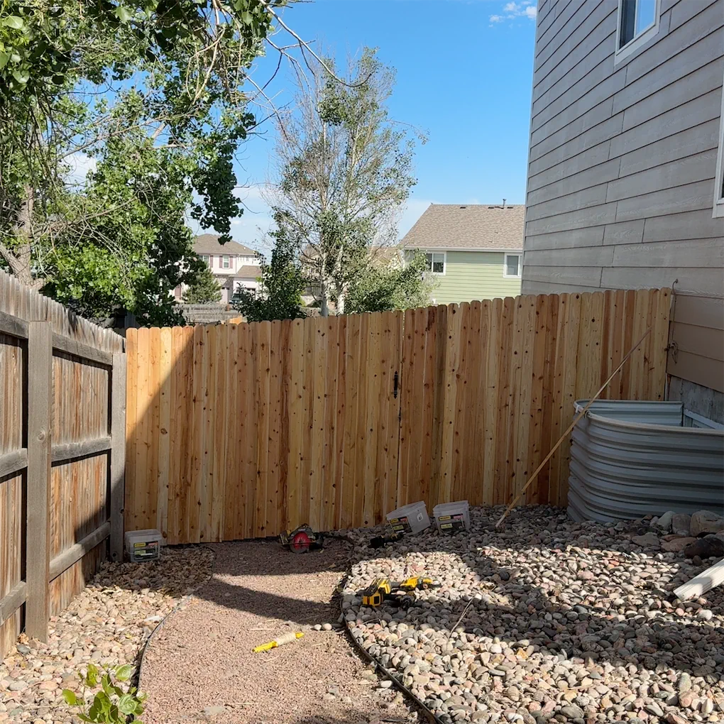 Backyard with a new wooden fence, some construction tools, and a gravel pathway. Trees and neighboring houses are visible in the background.