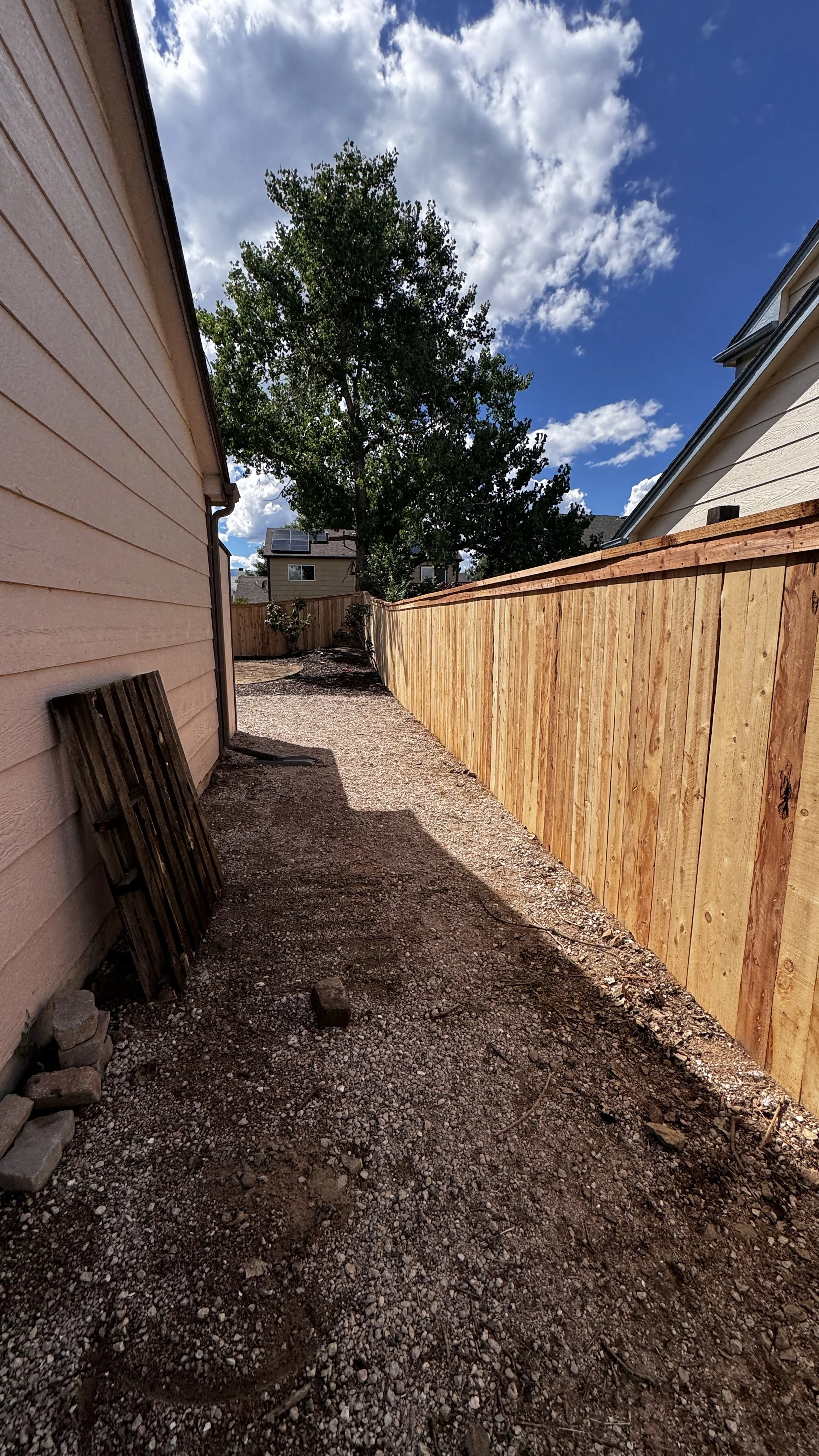 Side yard with a dirt and gravel ground, beige house on the left, new wooden fence on the right, and a large green tree in the background under a partly cloudy sky.