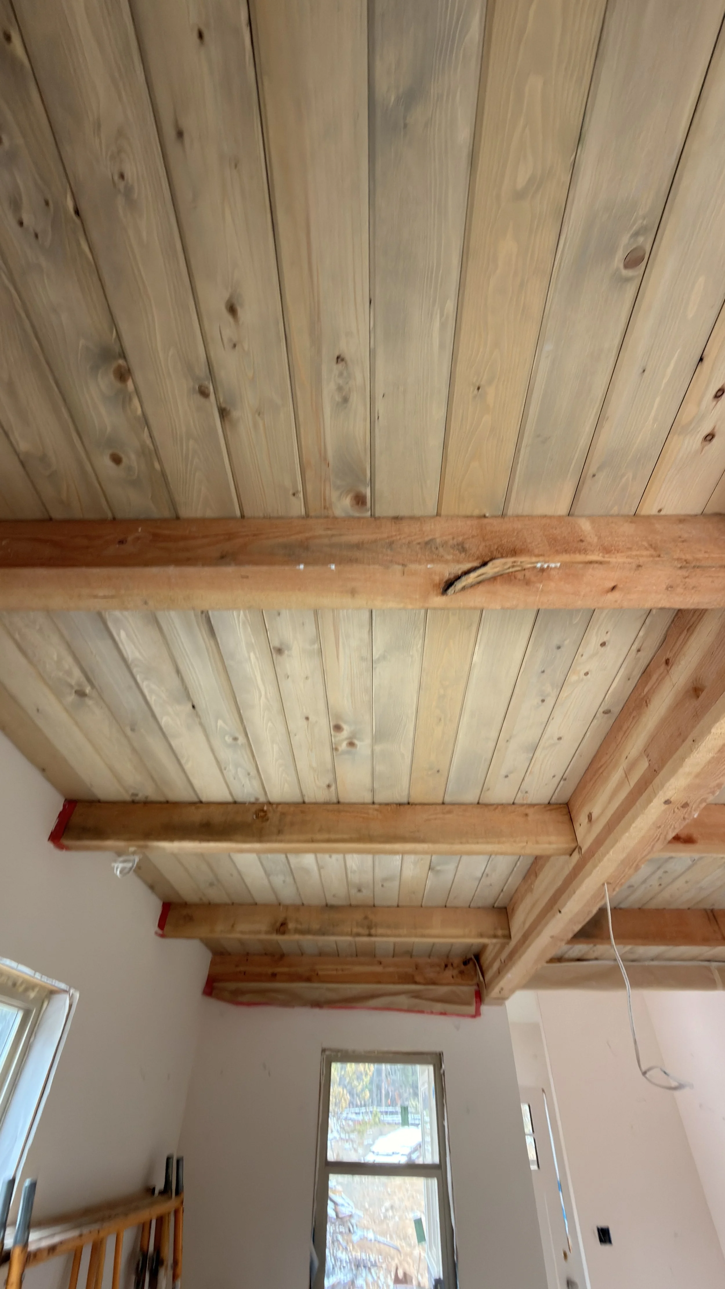 Interior view of a room under construction with a wooden ceiling and beams, two windows showing trees outside, and construction tools and wiring visible.
