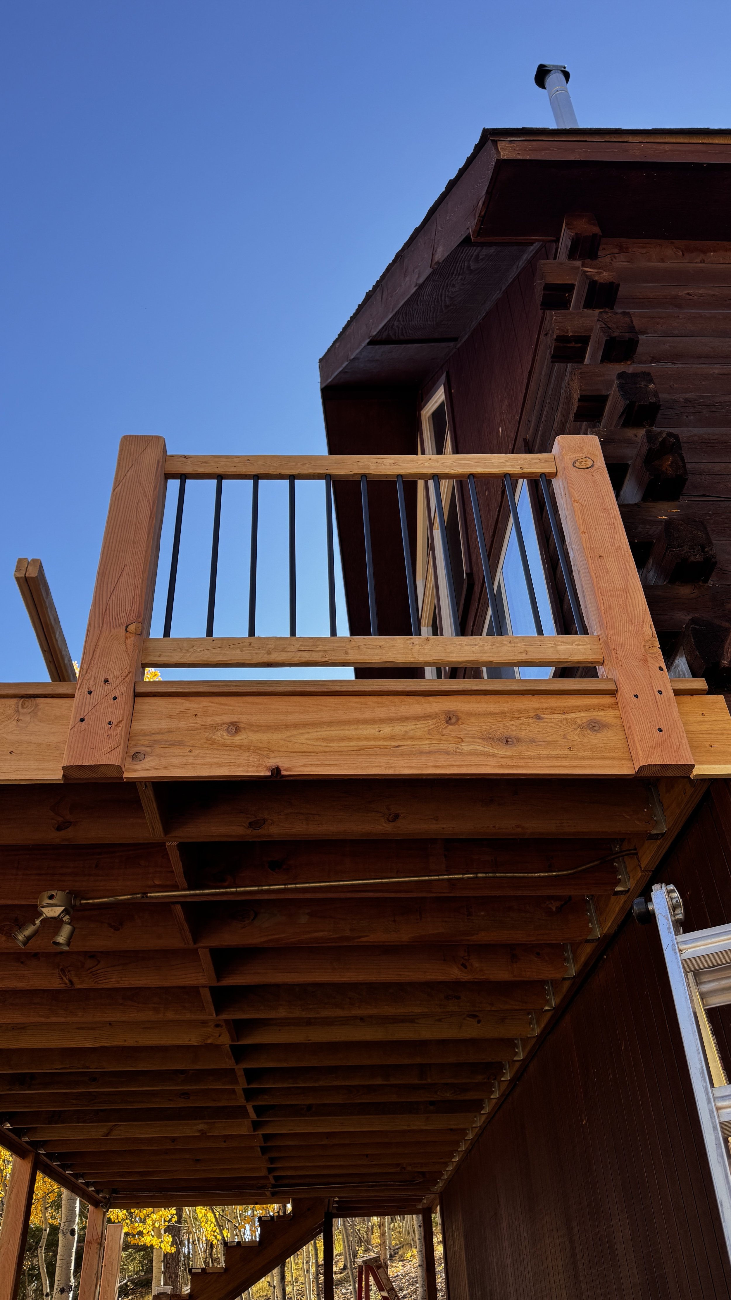 Photo of a wooden deck balcony with black metal railing and a house with dark brown wooden siding, under a clear blue sky. A ladder is leaning against the house, and trees with fall foliage are visible in the background.