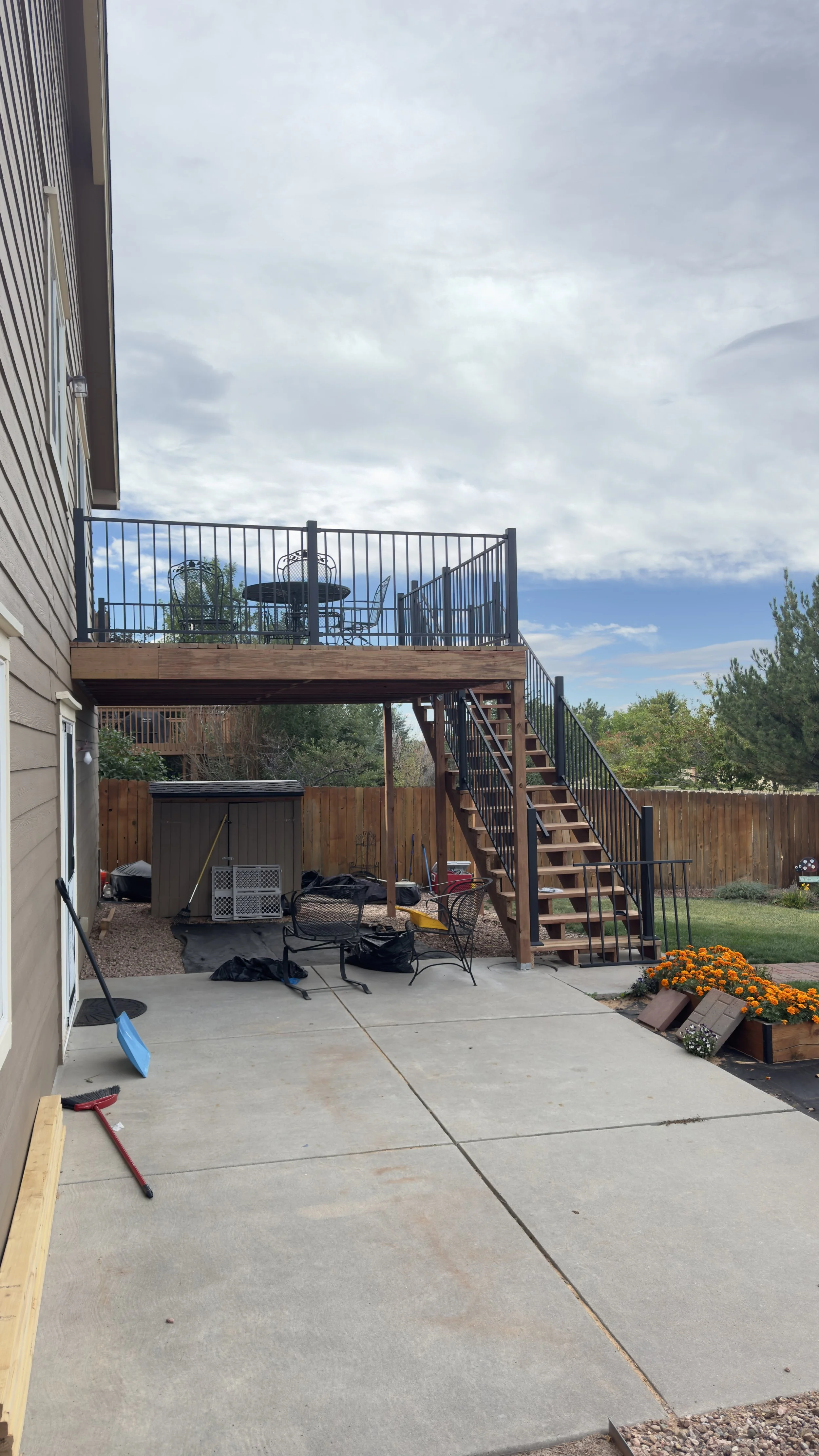 Backyard with a concrete patio, a two-story wooden deck with stairs, and a table and chairs on the upper deck. There are tools and construction materials scattered on the patio, a yard with a wooden fence, trees, a garden shed, and a flower bed with 
