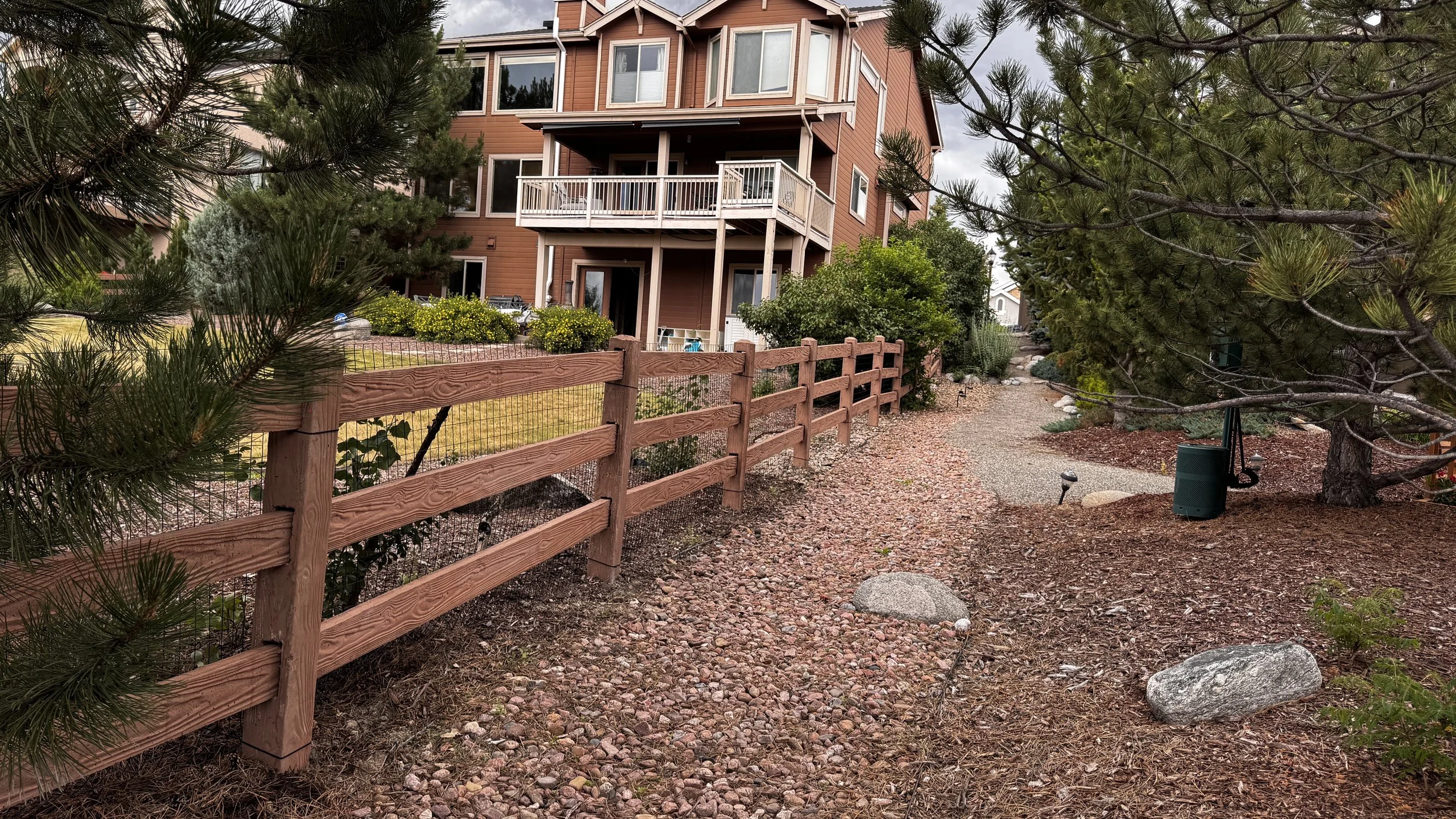 A multi-story house with a wooden exterior and multiple balconies, surrounded by trees and bushes, with a gravel and dirt pathway in the foreground and a green trash bin underneath a tree.