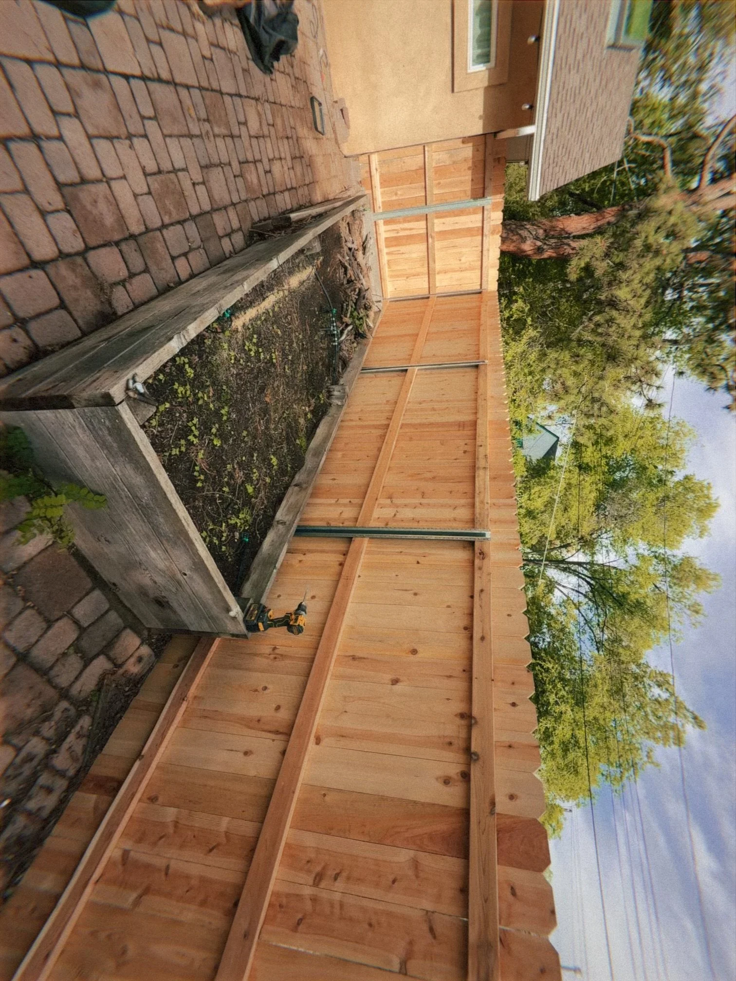 A backyard with a wooden fence, trees with green foliage, and a brick path with a raised garden bed. There is a power drill on the ground, and construction materials like wood and screws nearby. Part of a house with a window and shingle roof can be s