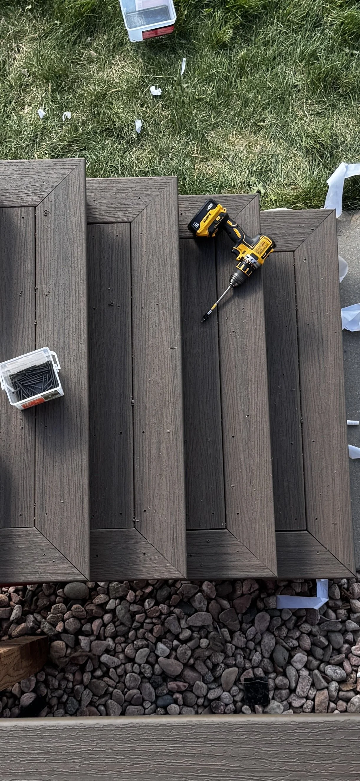 Close-up of a wooden deck with a cordless drill resting on the planks, a box of nails, and a container of screws nearby. The deck is above a layer of rocks, and grass is visible in the background.