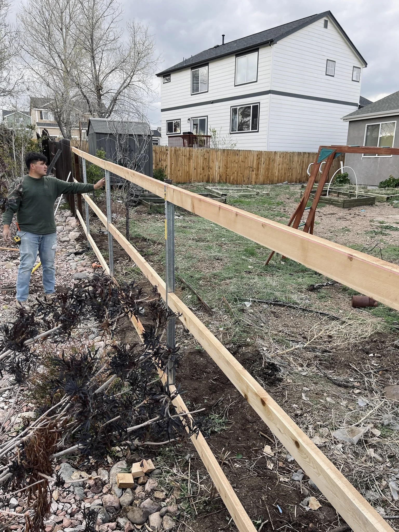 A person working on a construction project in a yard, installing a wooden fence. The yard has some plants, rocks, and a swing set frame, with a white house in the background under a cloudy sky.
