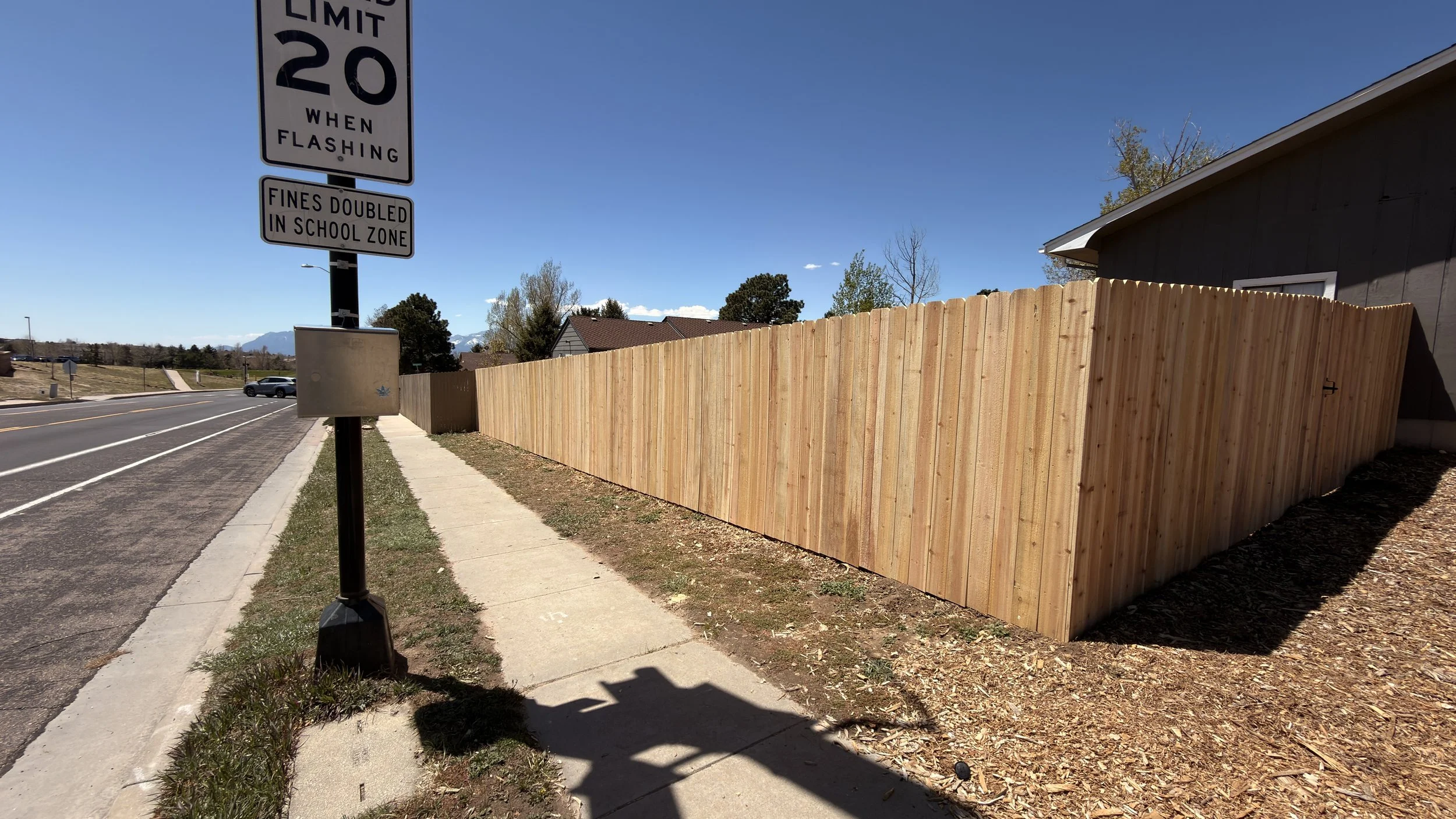 Street view with a sidewalk, a sign indicating a 20 mph speed limit when flashing, and a tall wooden fence running along the side of a house. Mountains can be seen in the background under a clear blue sky.