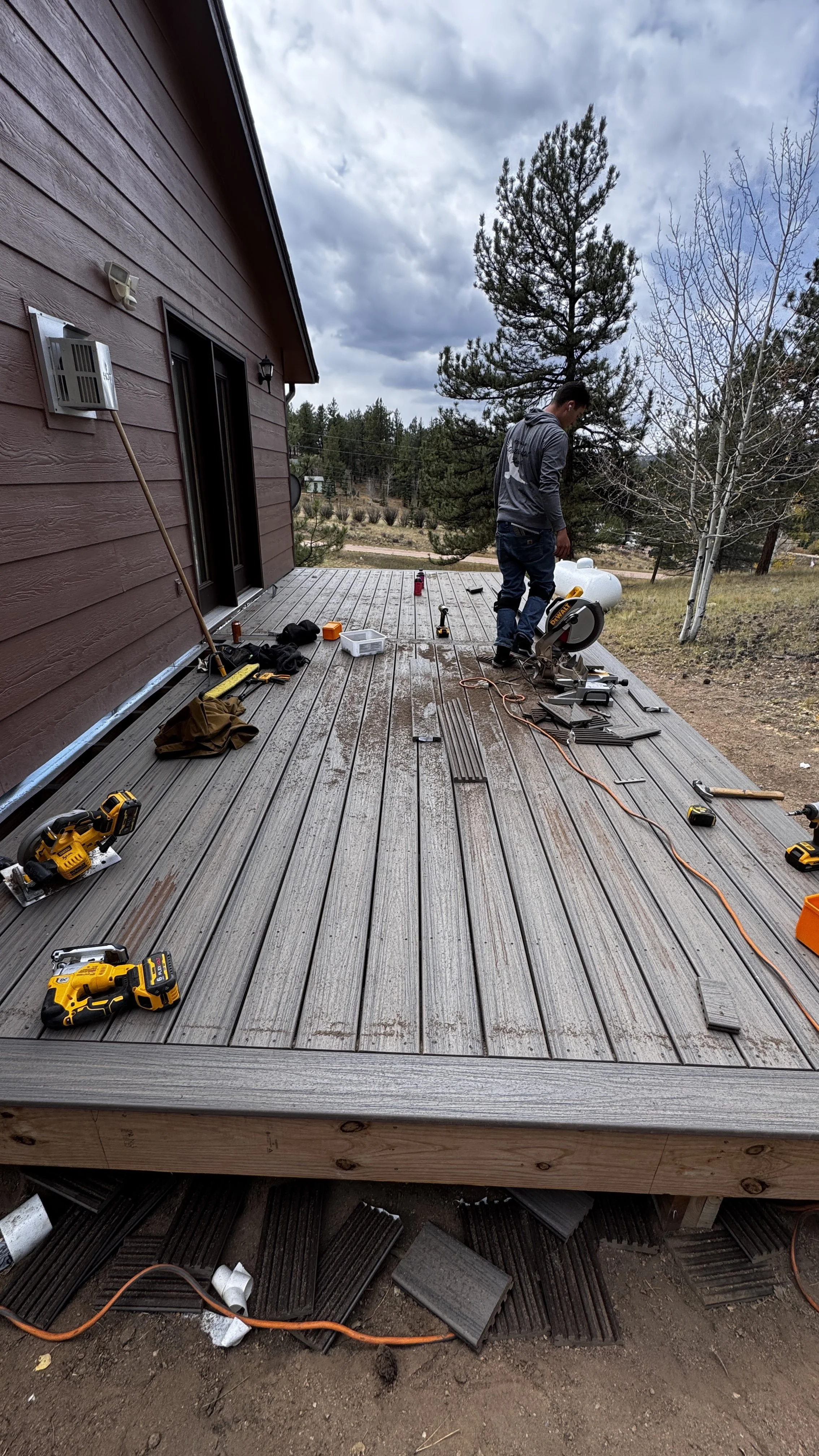 Man installing a wooden deck outside house on cloudy day. Tools and materials, including power saw, drill, and screws, spread on the deck.