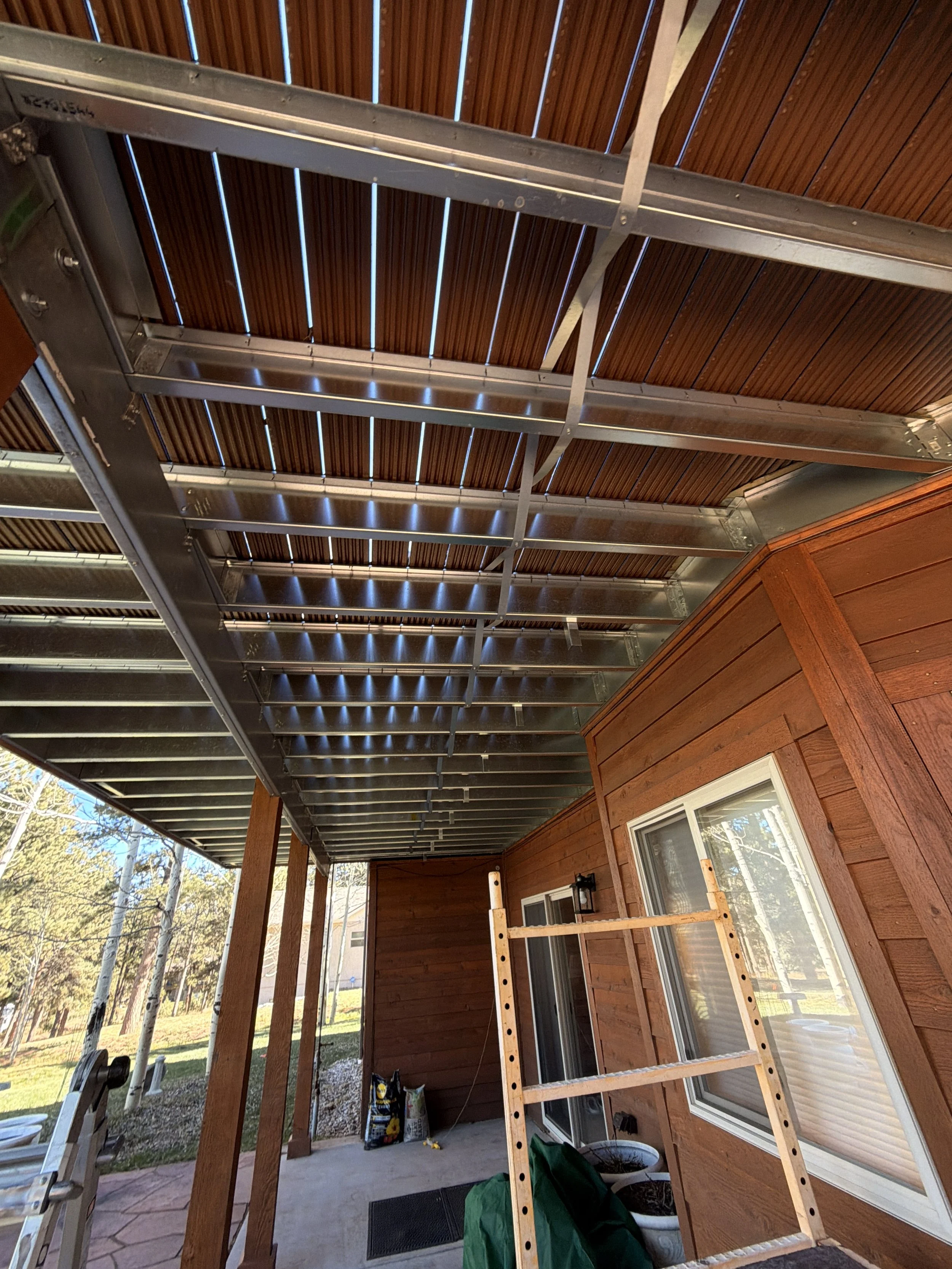 Back porch area with a wooden overhang, ladder, construction supplies, and a wooden house exterior.