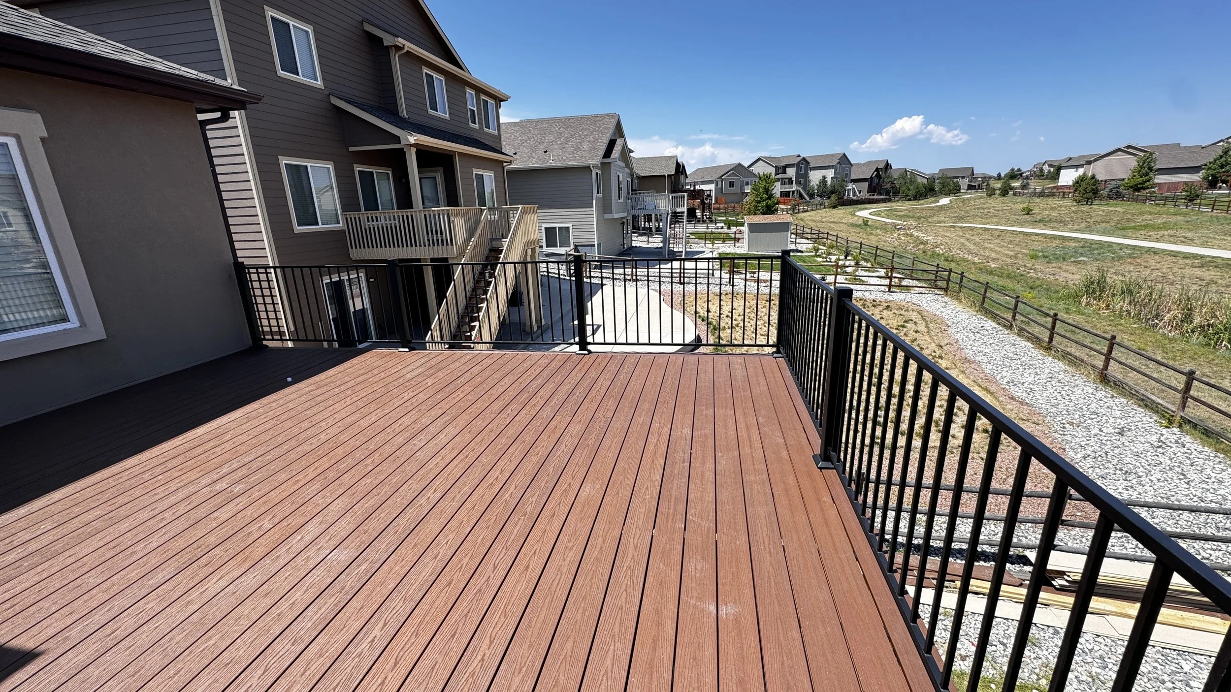 View from a wooden deck overlooking a residential neighborhood with multiple houses, a gravel path, a grassy area, and a clear blue sky.