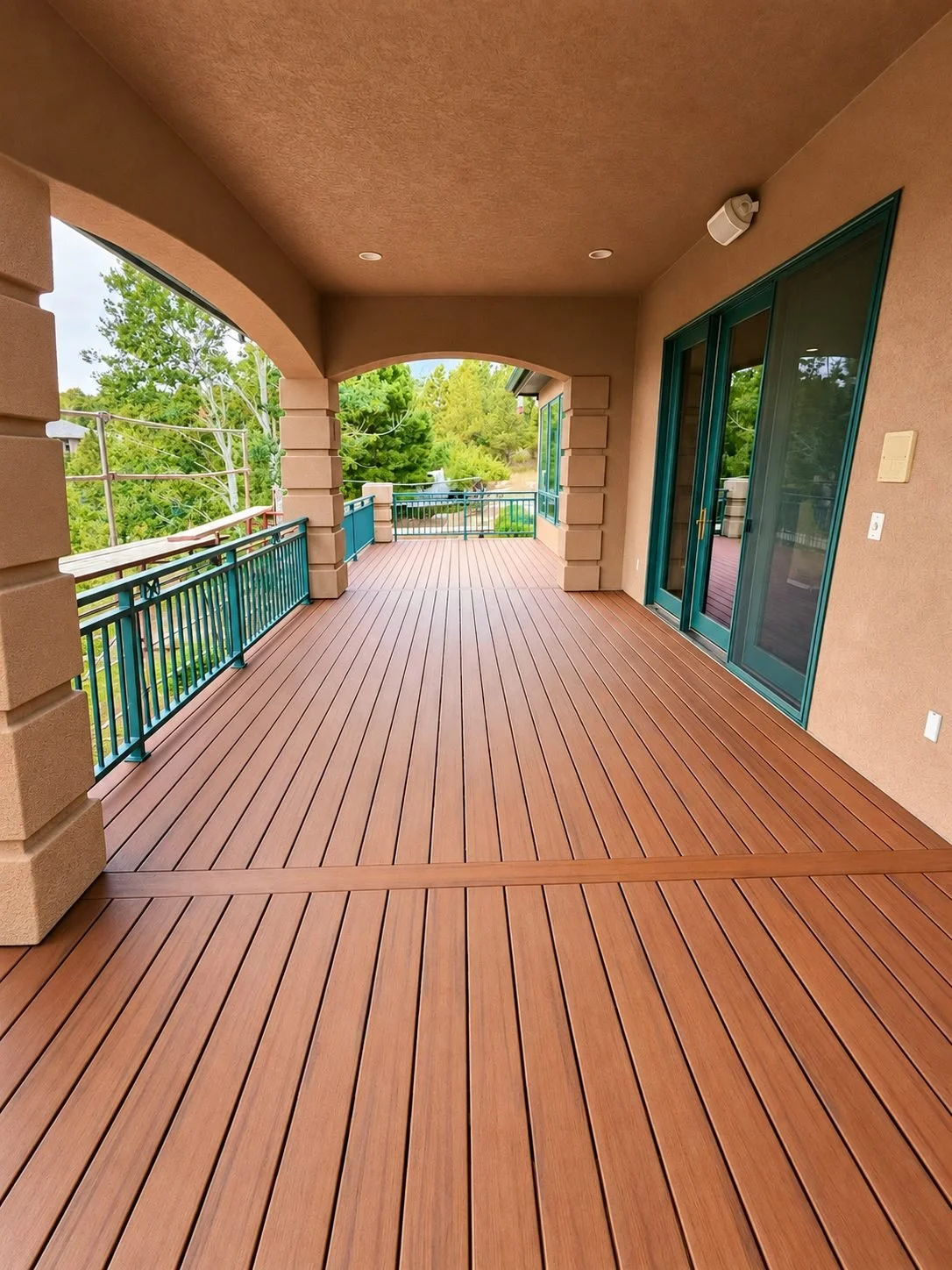 Covered deck with reddish-brown decking, teal metal railing and stucco arches, with trees and a mountain view beyond