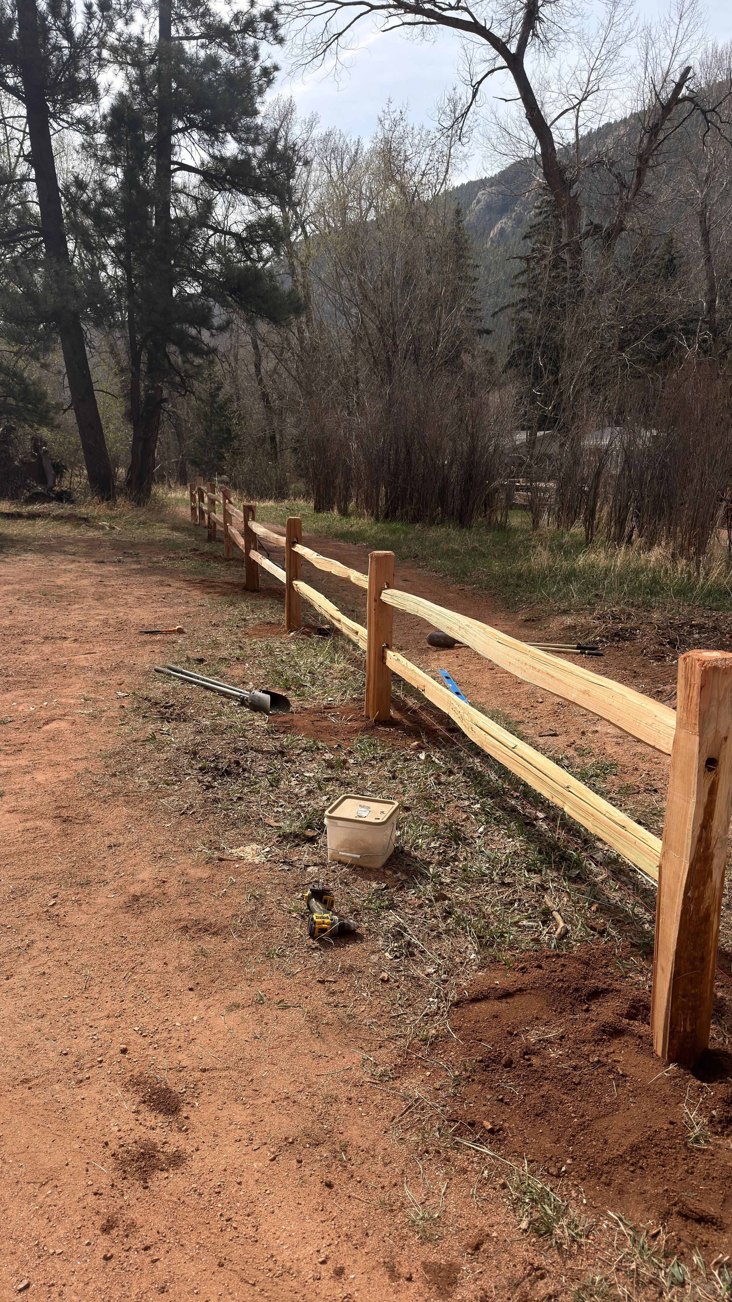 Wooden fence being built along a dirt path in a forested area with trees and mountains in the background.