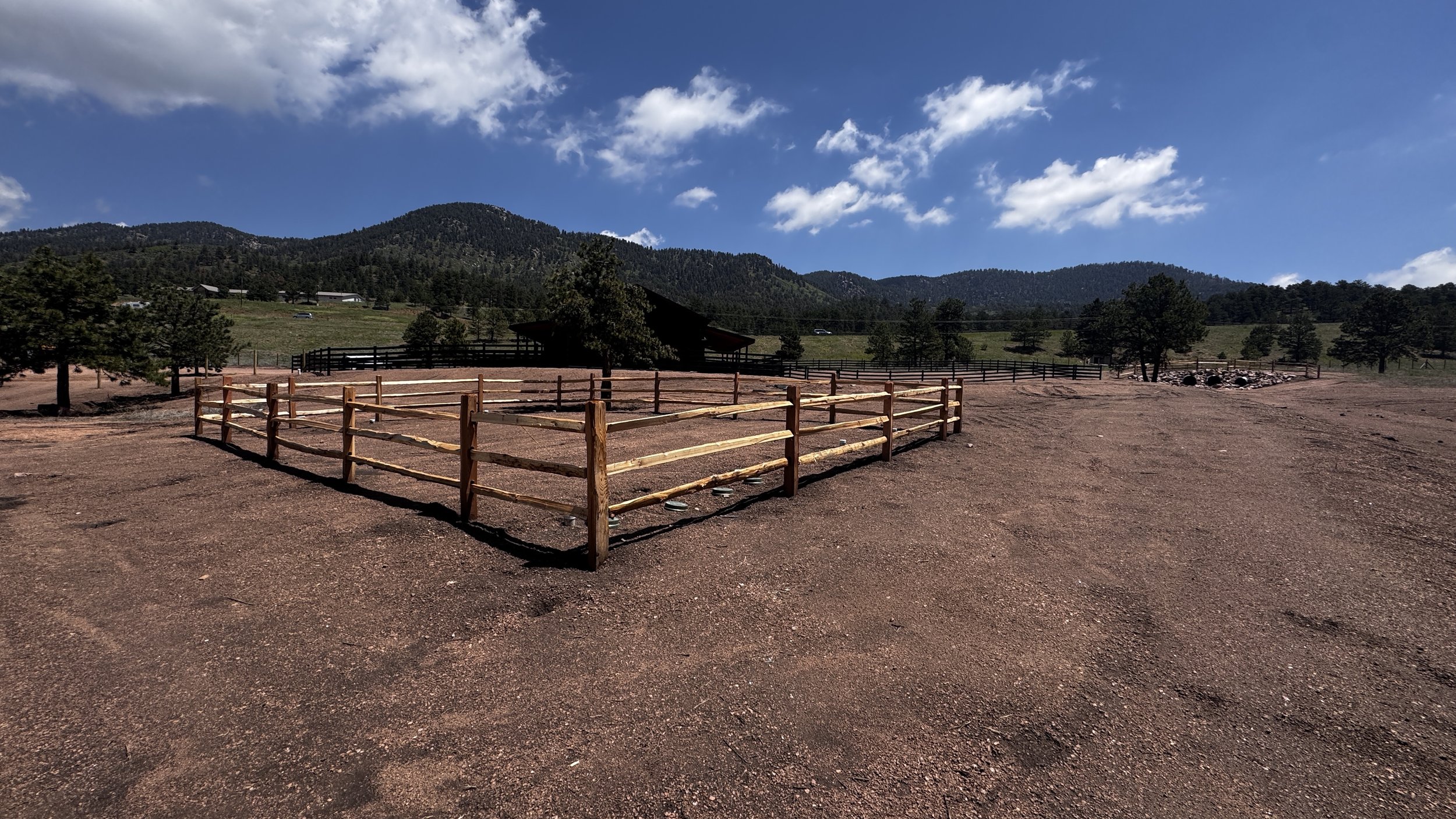 A fenced outdoor area with a reddish dirt ground, surrounded by trees and mountains under a partly cloudy blue sky.