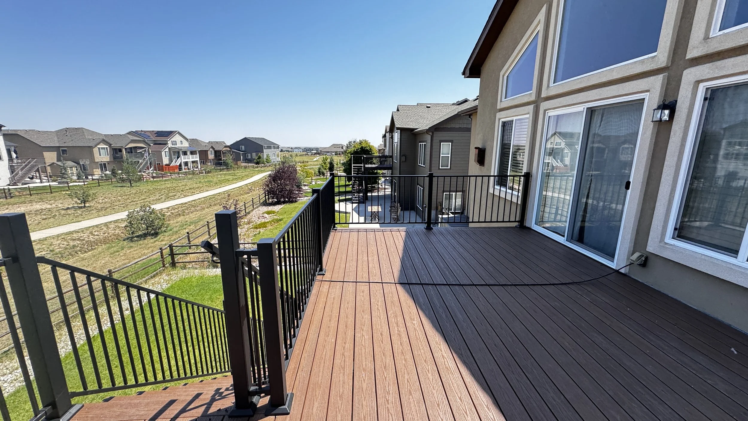 View from a balcony of a residential neighborhood with multiple houses, a walking path, and some trees, under a clear blue sky.