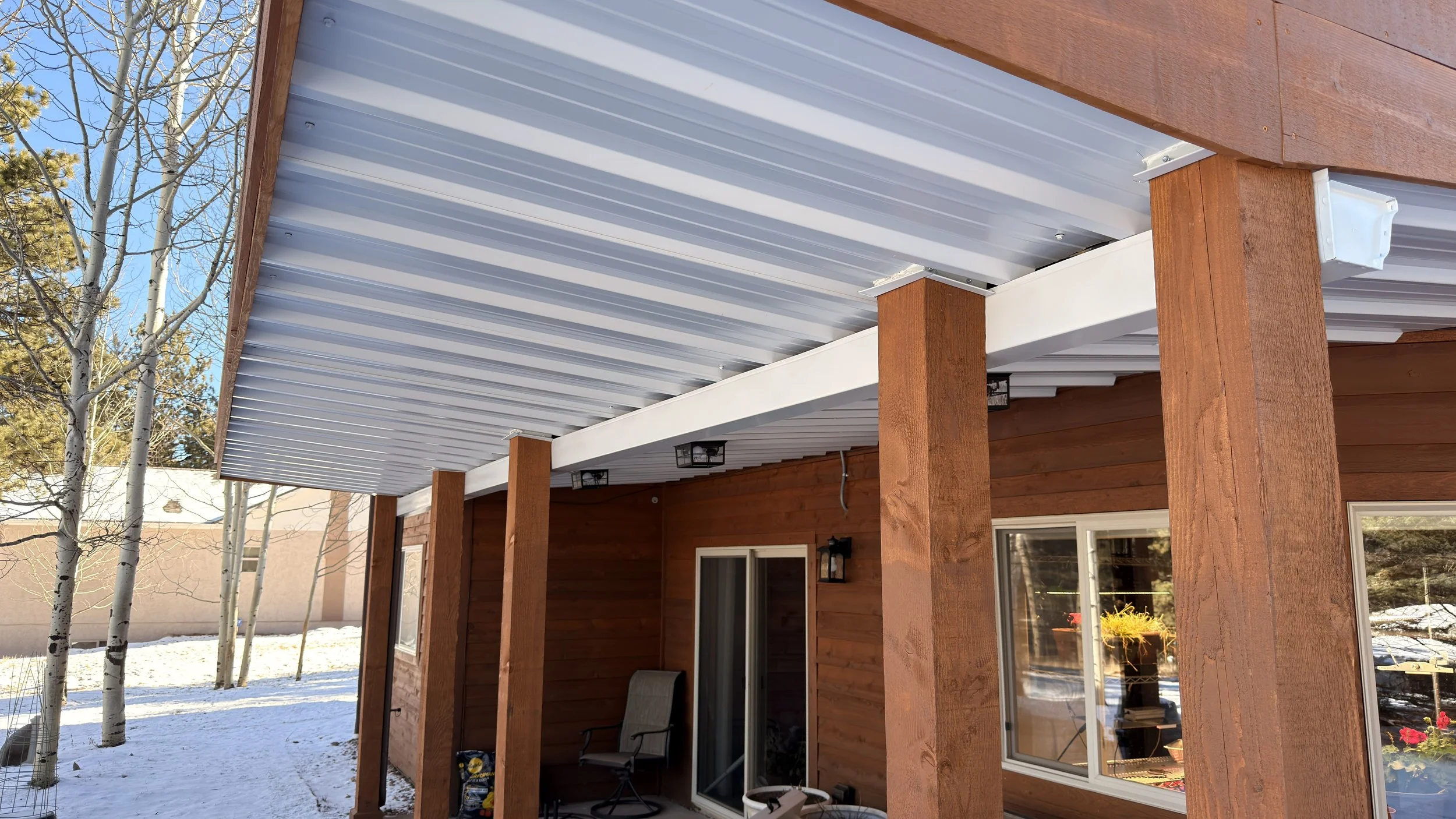 Backyard patio area with a corrugated metal roof supported by wooden posts, attached to a wooden house. There are leafless trees and snow on the ground, with a clear blue sky.
