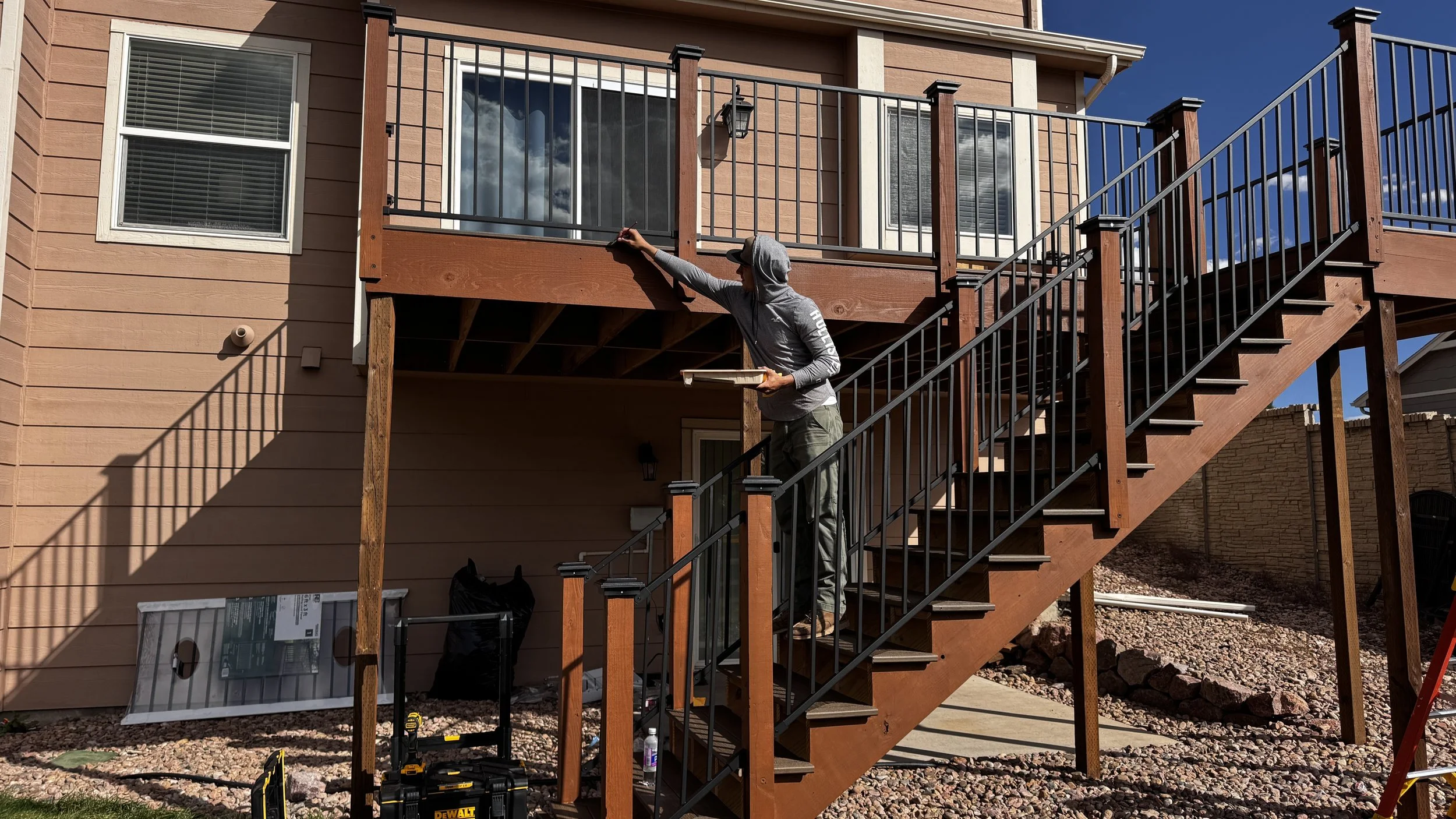 Person wearing a hoodie painting or staining a wooden balcony railing on a second-story deck of a house, with stairs leading up to it. Construction tools and materials are visible below.