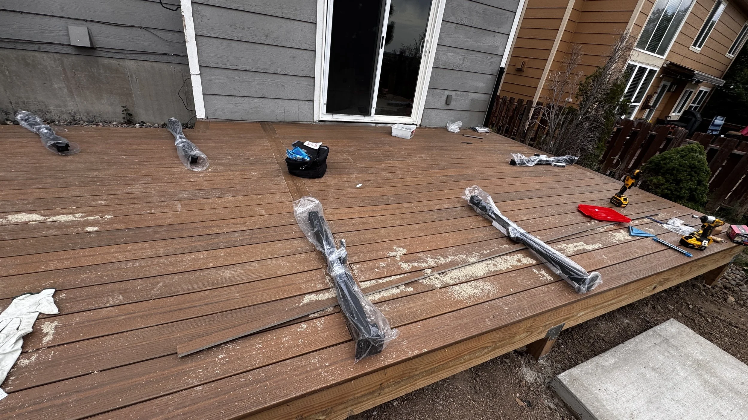 A wooden deck under construction with tools and supplies scattered on it, including drills, a red dustpan, plastic wrap, and small bags, in front of a house with gray and tan exterior walls and a sliding glass door.