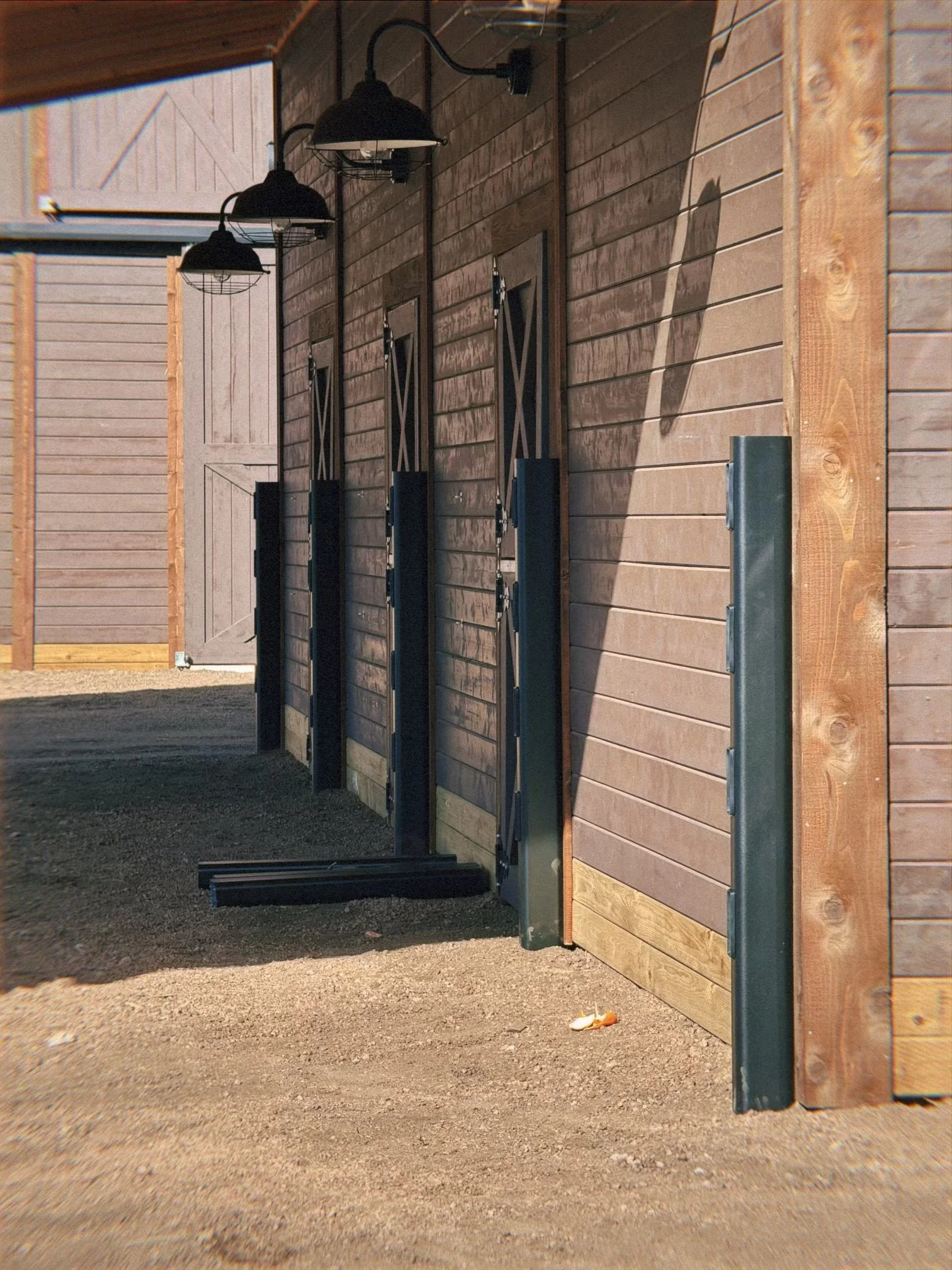 Close-up of wooden exterior wall of a stable with three black hanging lights, metal brackets, and a gray door in the background. A dirt ground and a discarded cup are visible on the ground.