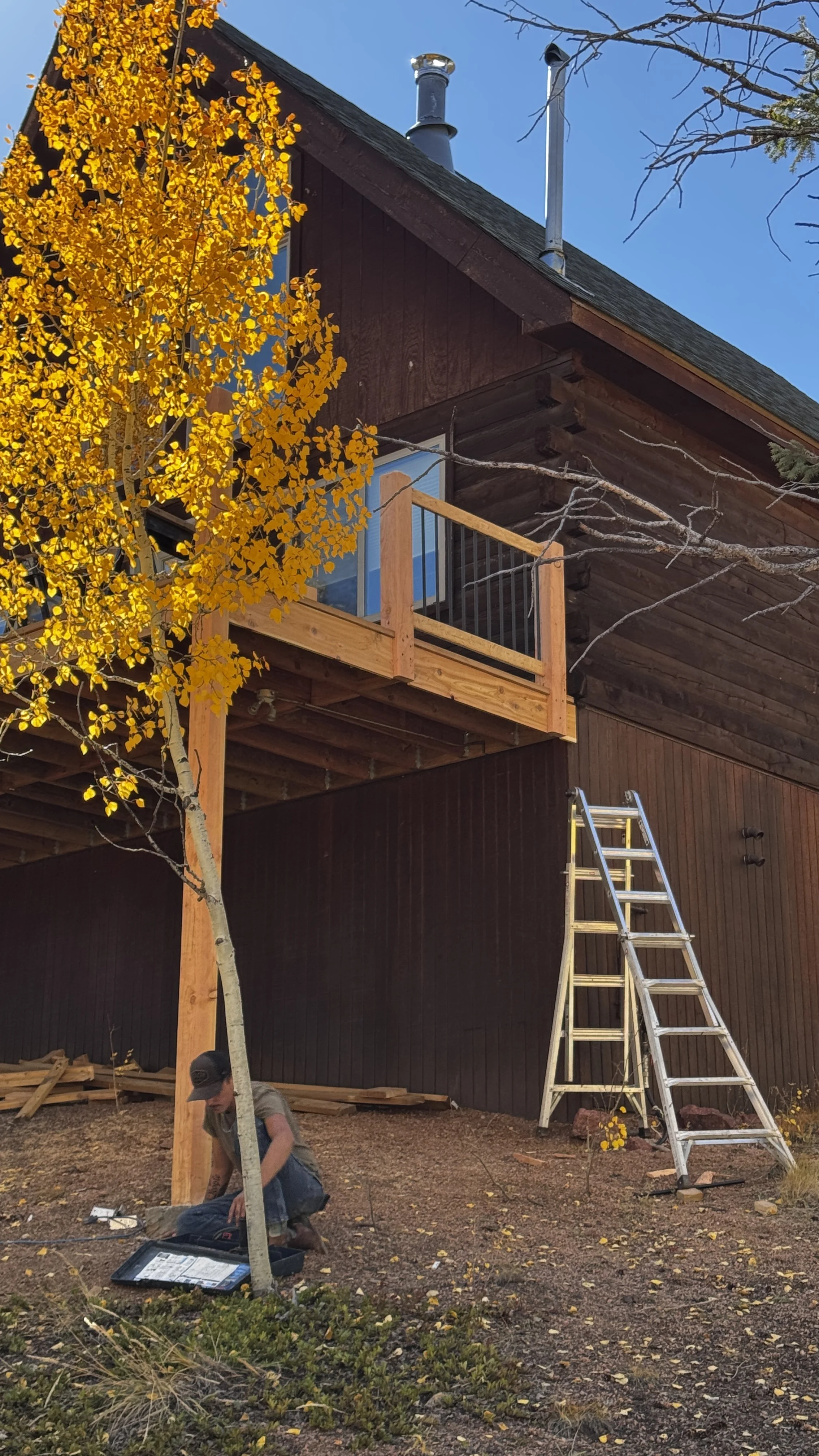 A person working on a construction project outside a brown wooden house with a small balcony. A metal ladder is leaning against the house, and a yellow-leaved tree is in the foreground. The sky is clear and blue.