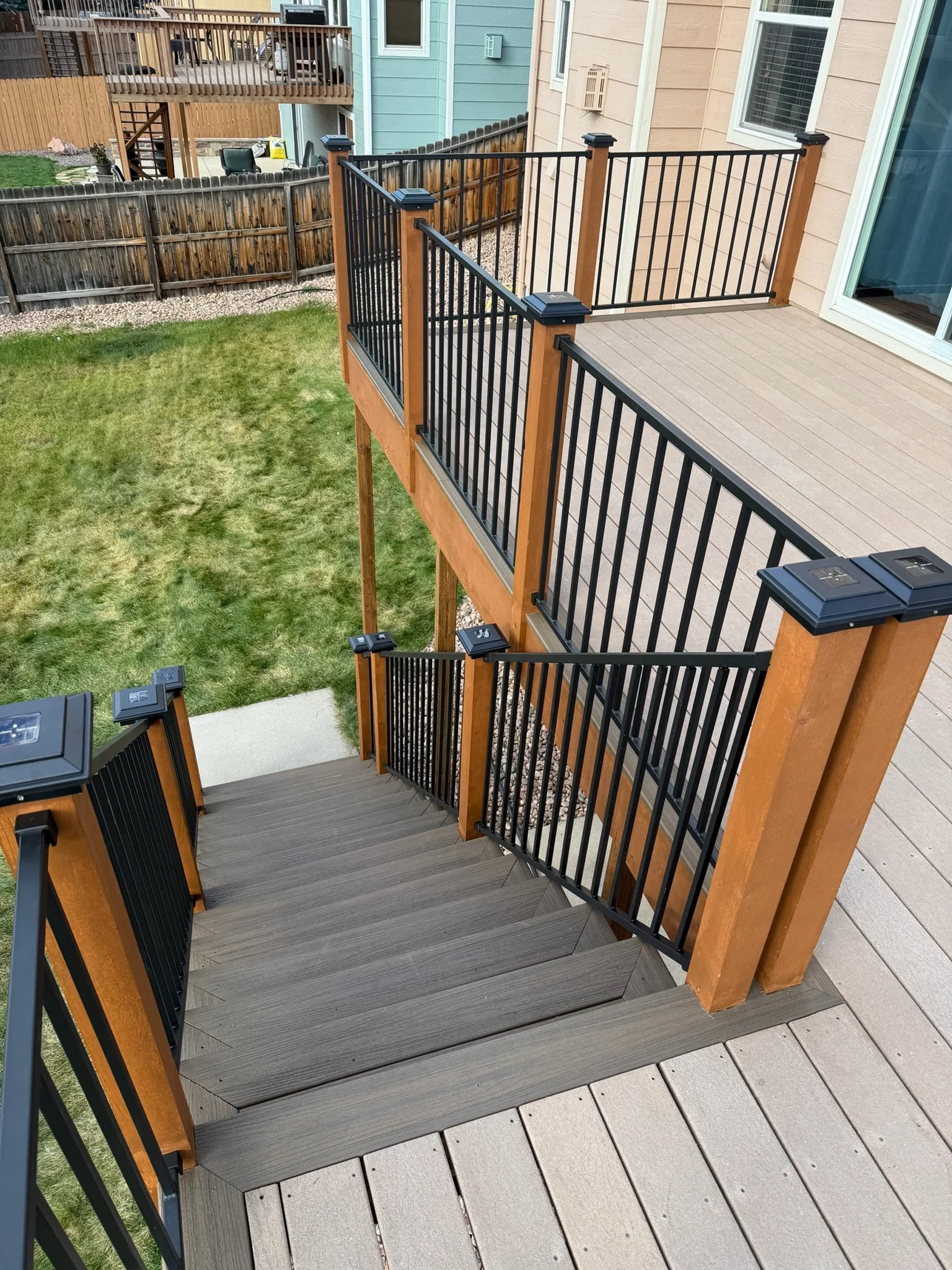 View of a backyard deck with railing, stairs, and grass yard, adjacent to neighboring houses with fences.