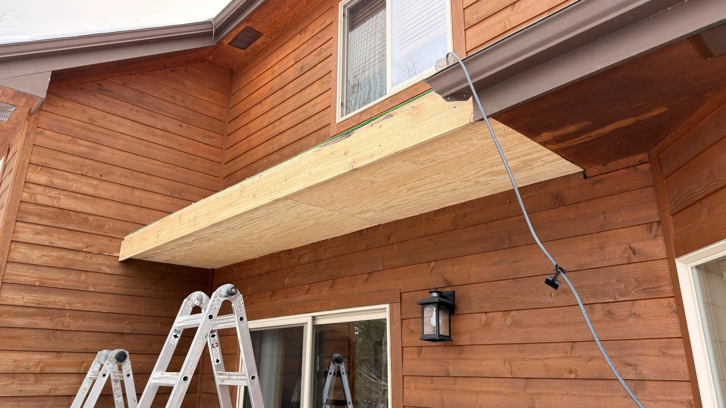 Unfinished wooden balcony extension on a house with red-brown siding, a sliding glass door, and a black outdoor lantern. A ladder and a work light are present.