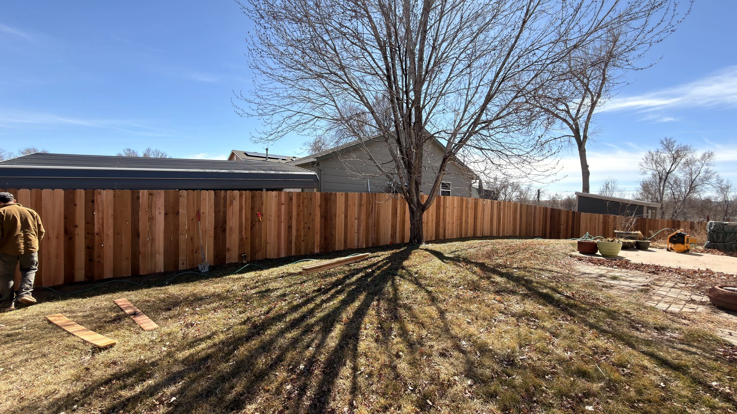 Backyard with a wooden fence, leafless tree, patio area with potted plants, garden tools, and lawn equipment. Partial view of a person in a brown jacket on the left. Clear blue sky.