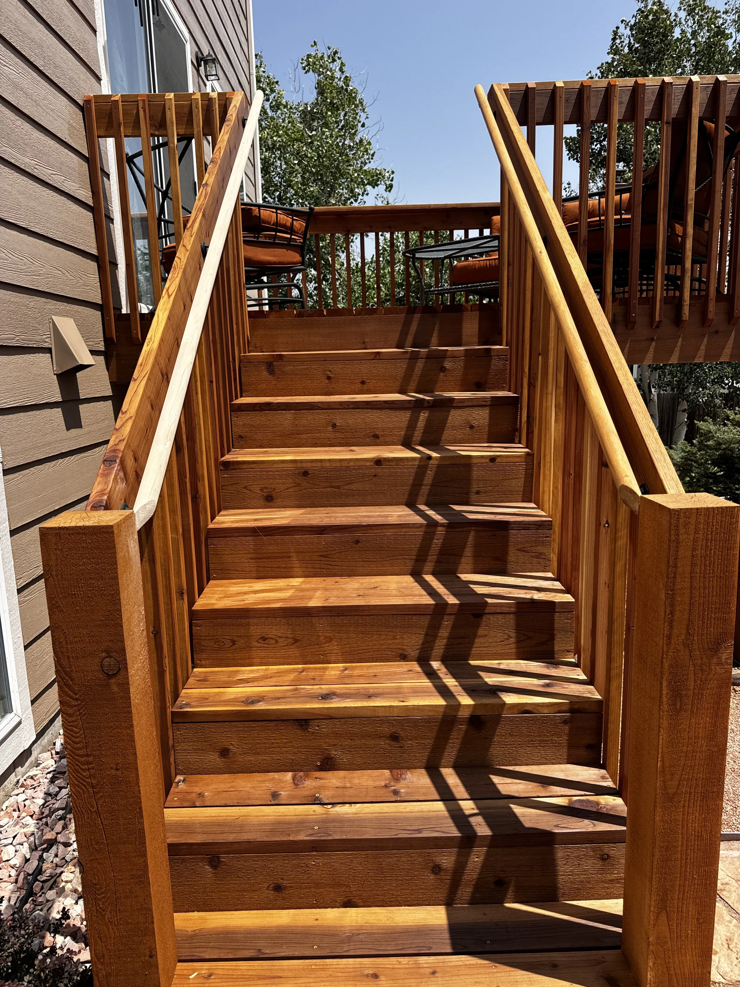 A wooden staircase leading to an outdoor deck with a table and chairs, attached to a house with beige siding and a window, with trees and clear blue sky in the background.