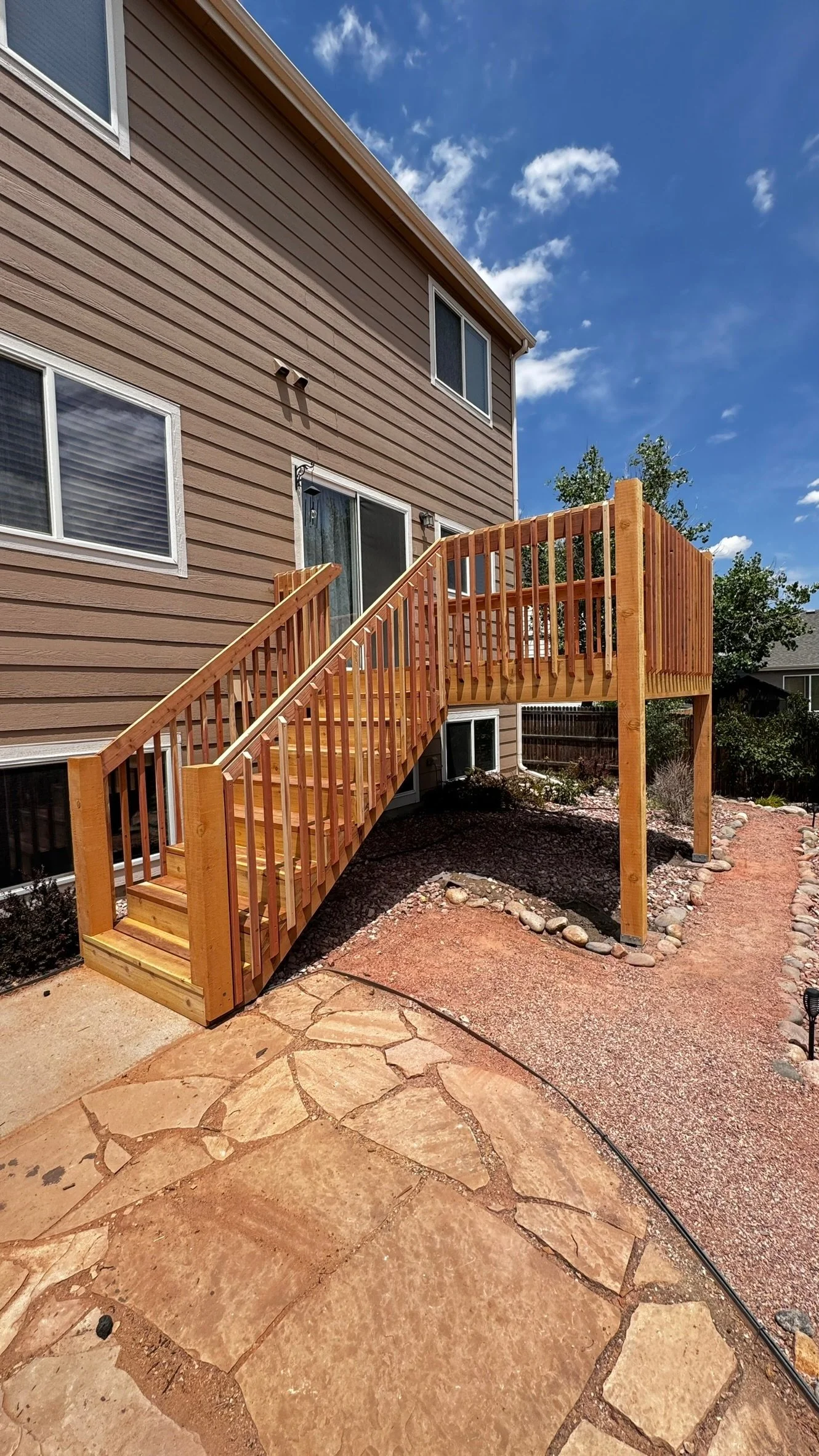 A newly built wooden deck with stairs attached to the back of a beige two-story house with white-framed windows. The deck has a railing around it and leads to a backyard area with rocks, gravel, and a tree. The sky is partly cloudy.