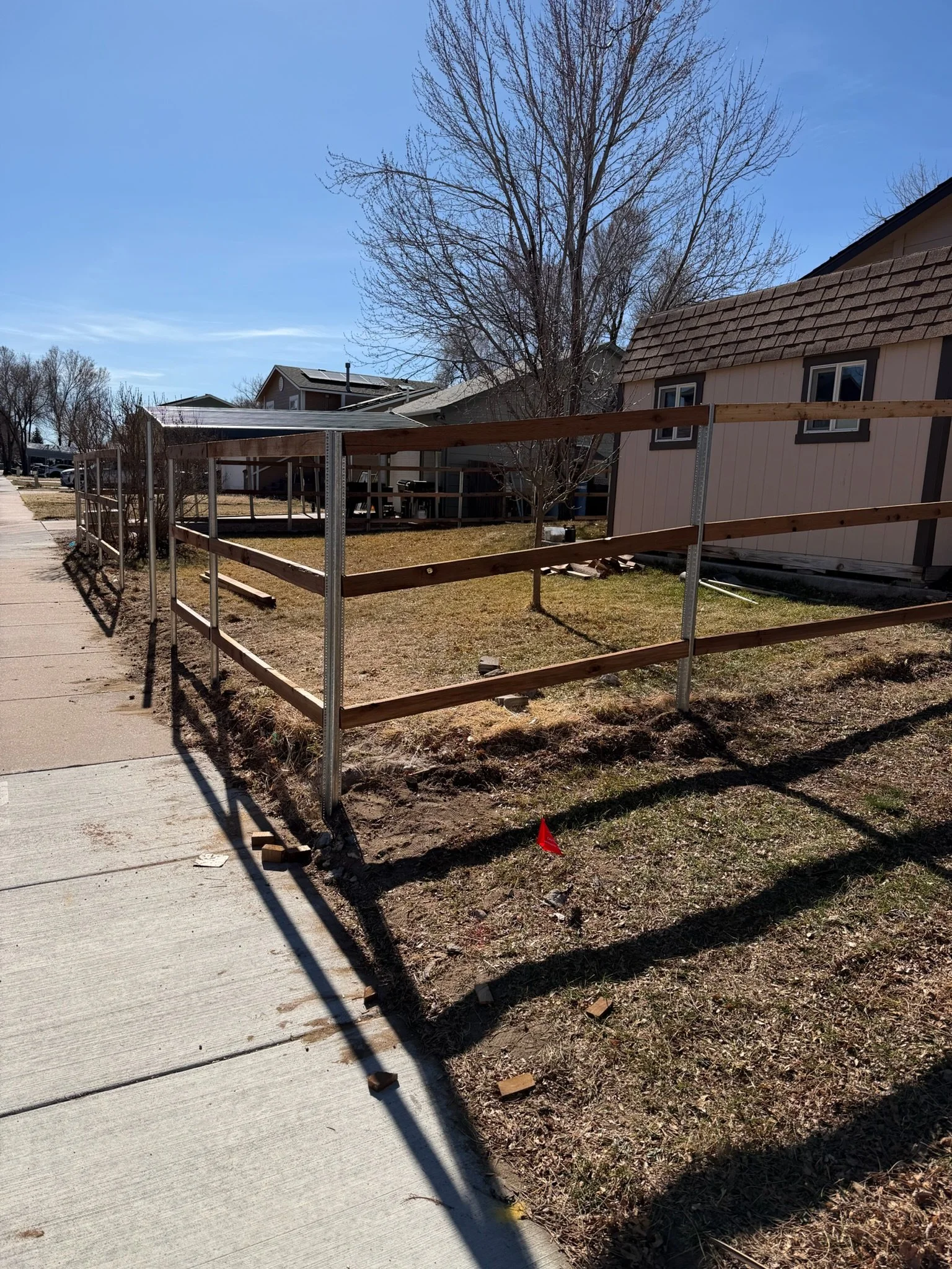 A wooden fence under construction in a backyard, with a house, leafless tree, and clear blue sky in the background.