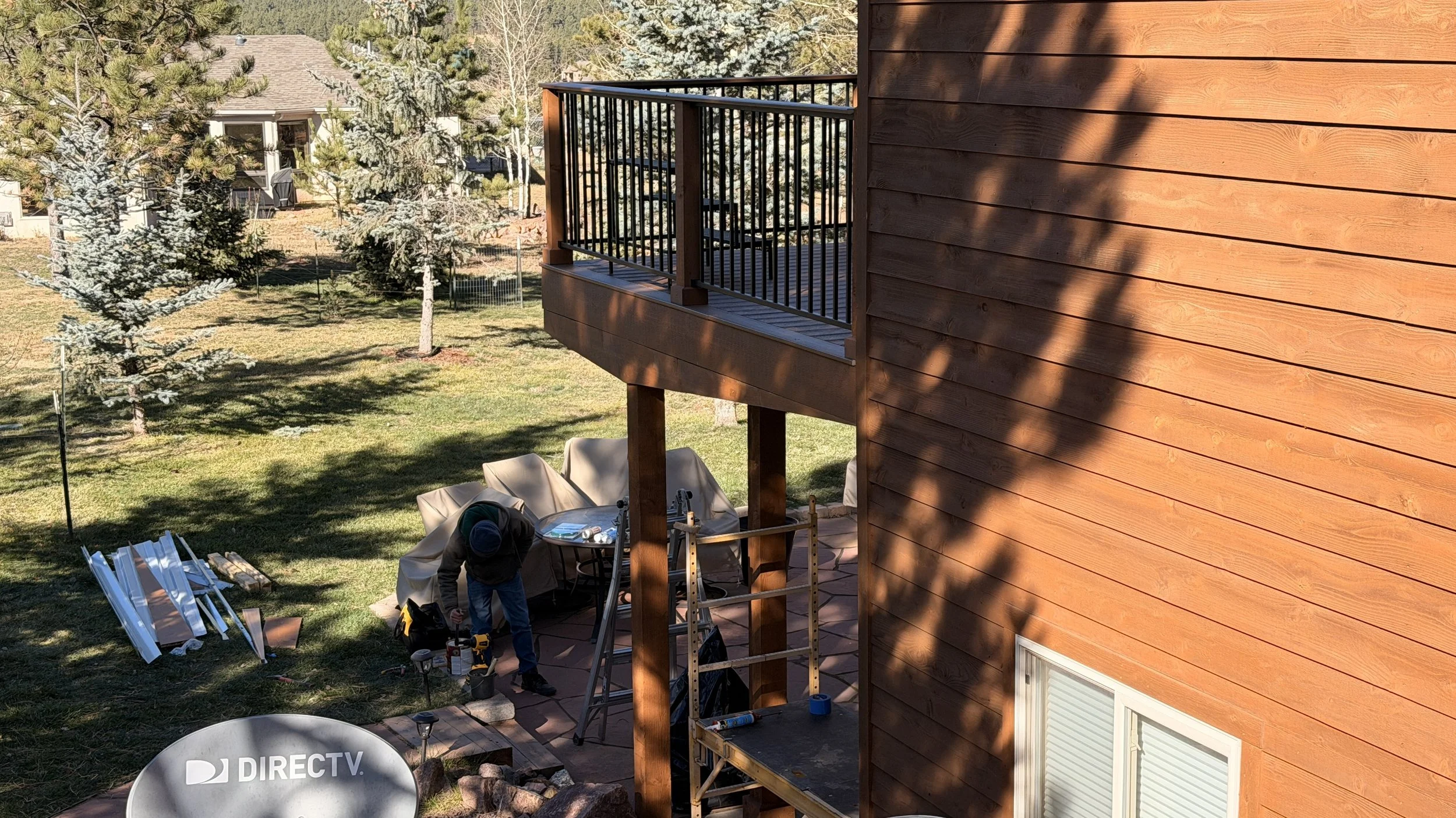 A worker building a wooden deck on the second floor of a house, surrounded by trees and a backyard with construction materials and tools.