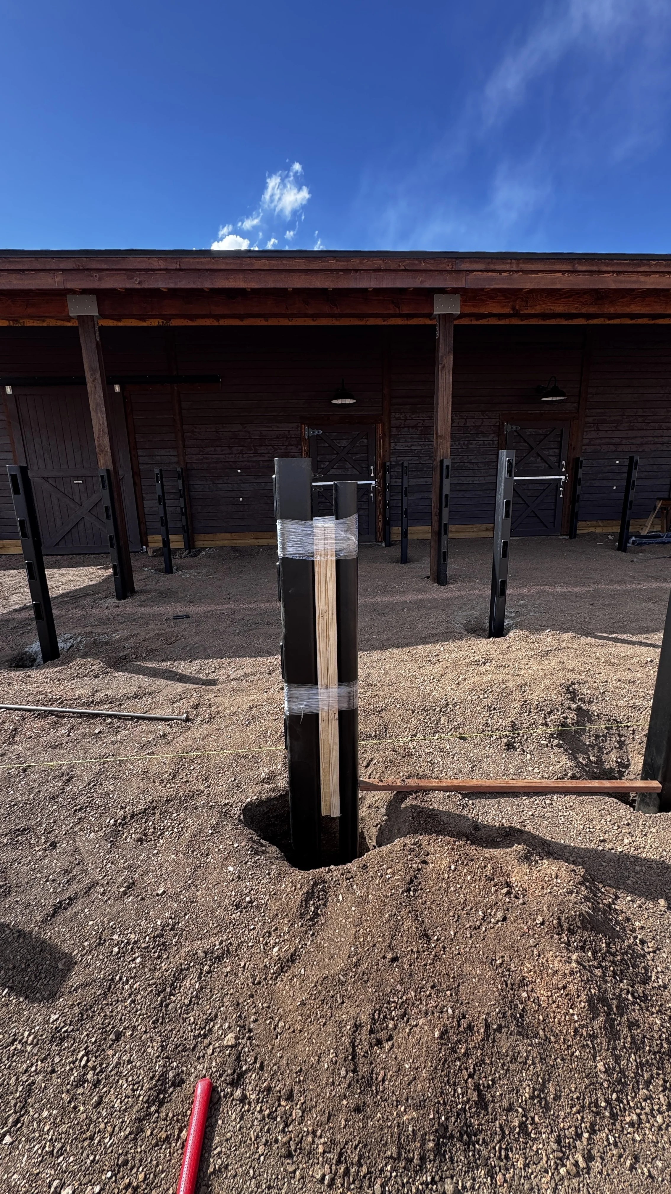 Construction site with a wooden building and metal poles in the dirt, a wooden post wrapped with plastic and a red hose on the ground.