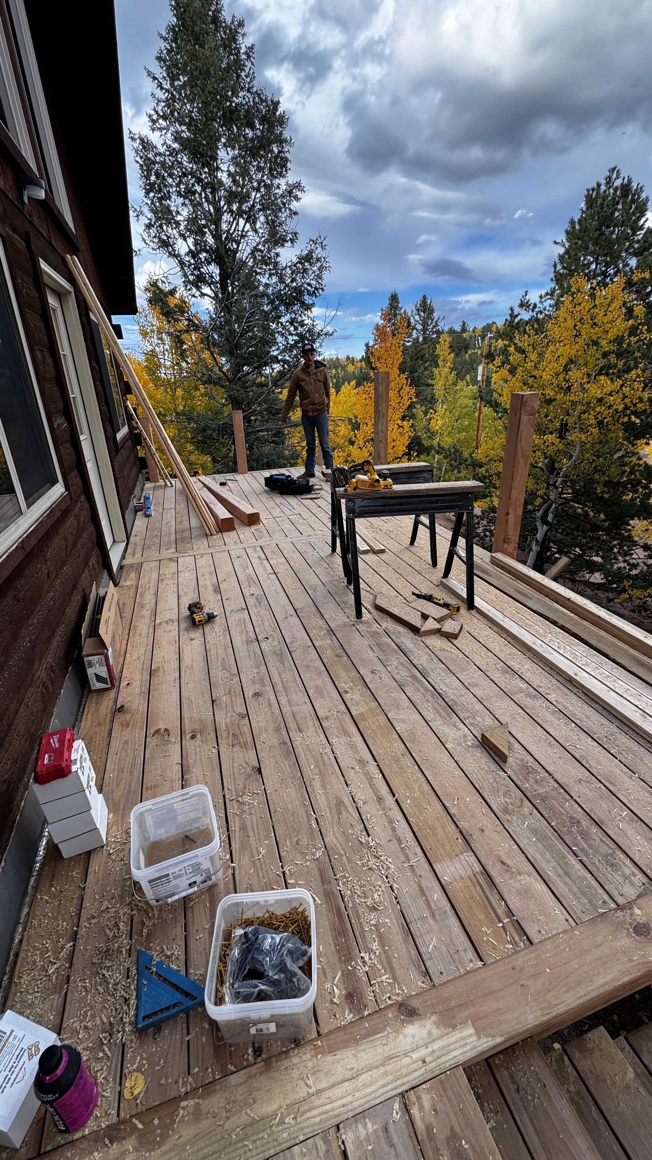 View of a decking project under construction on an outdoor wooden deck, with tools, construction materials, and a person standing among the work area, surrounded by trees with fall foliage and a cloudy sky.
