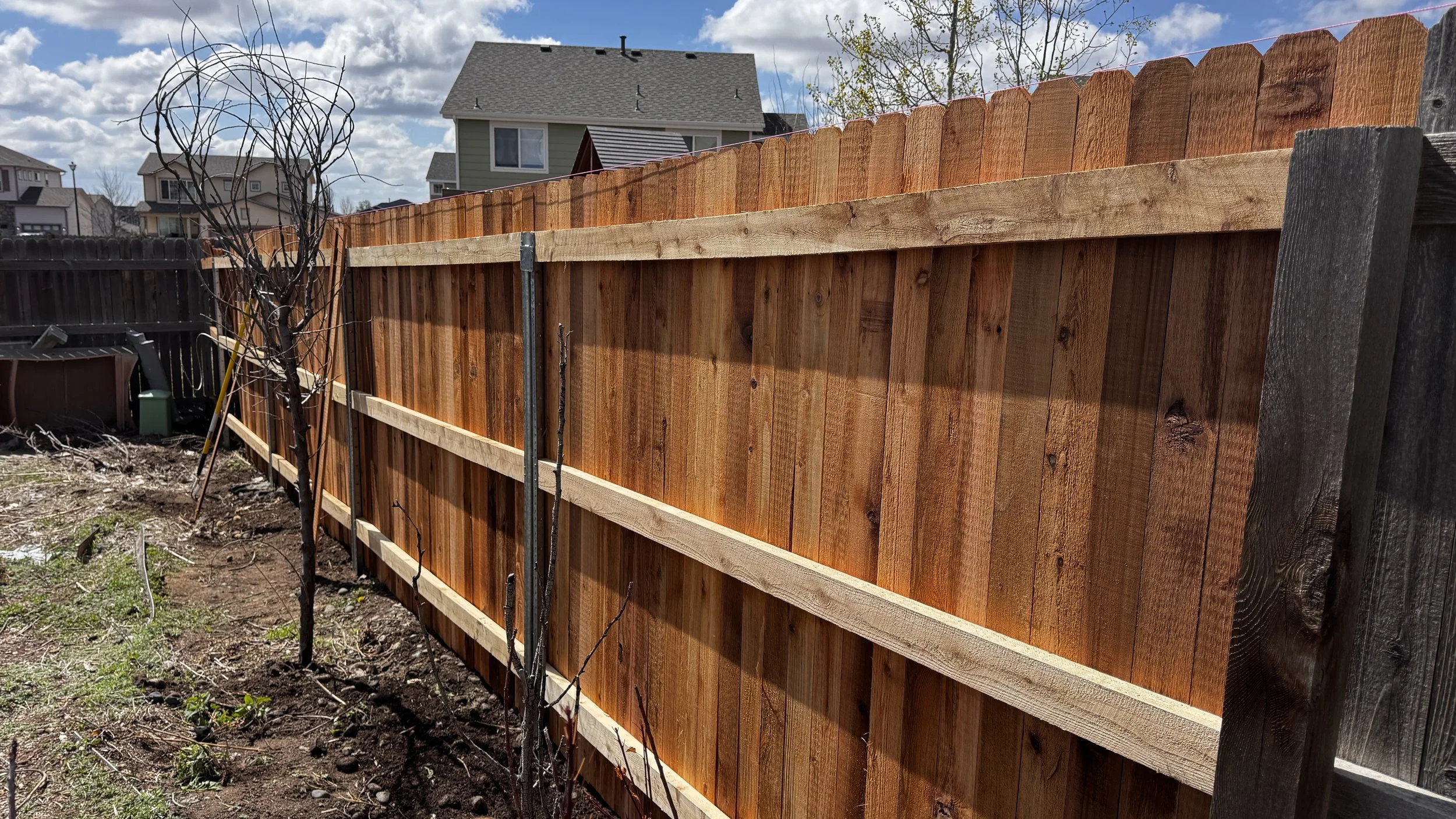 A newly installed wooden privacy fence with vertical planks surrounds a residential backyard, with houses and a partly cloudy sky in the background.