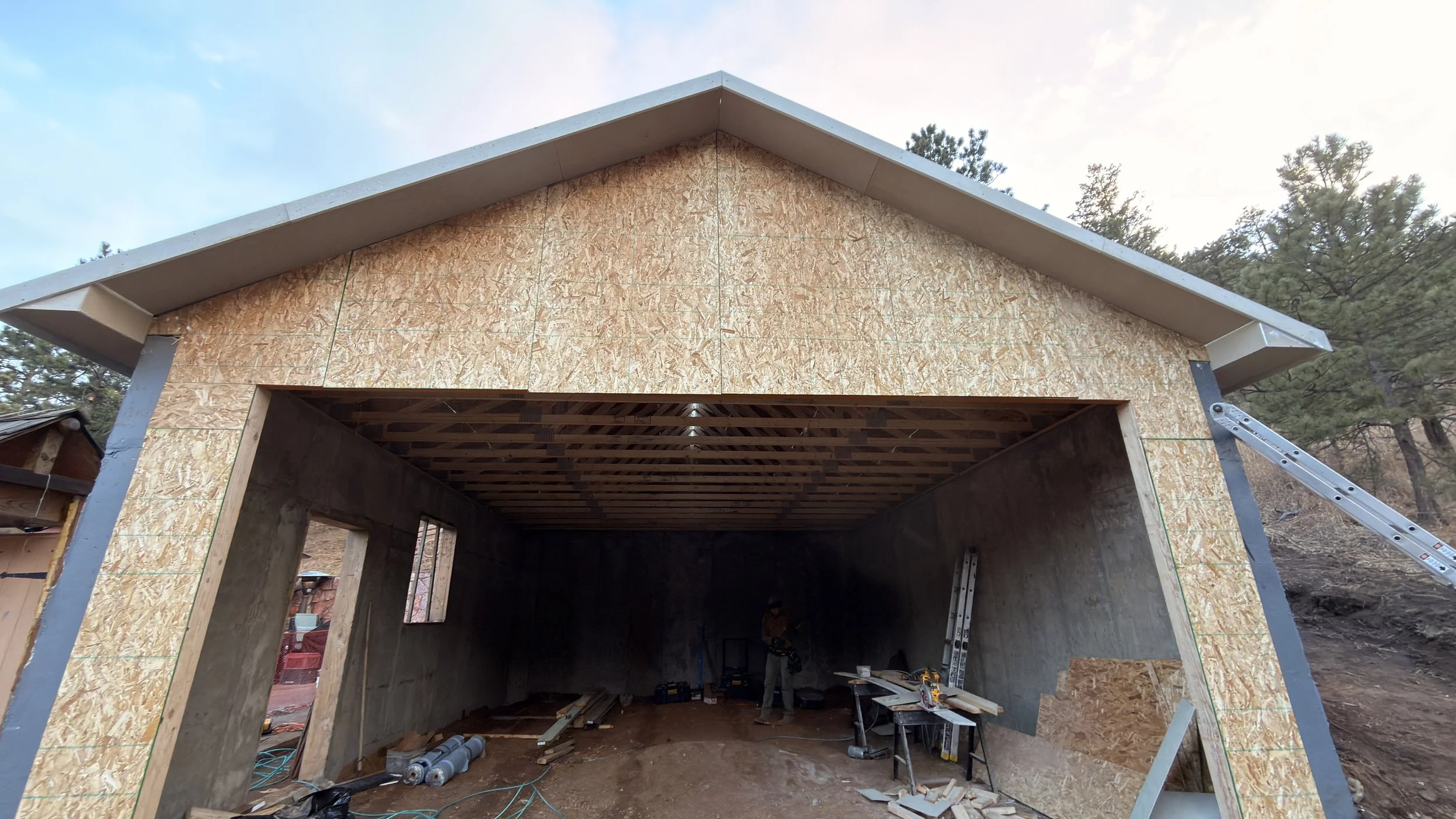 A building under construction with an exposed wooden frame and OSB sheathing on the exterior walls, surrounded by trees and construction tools.